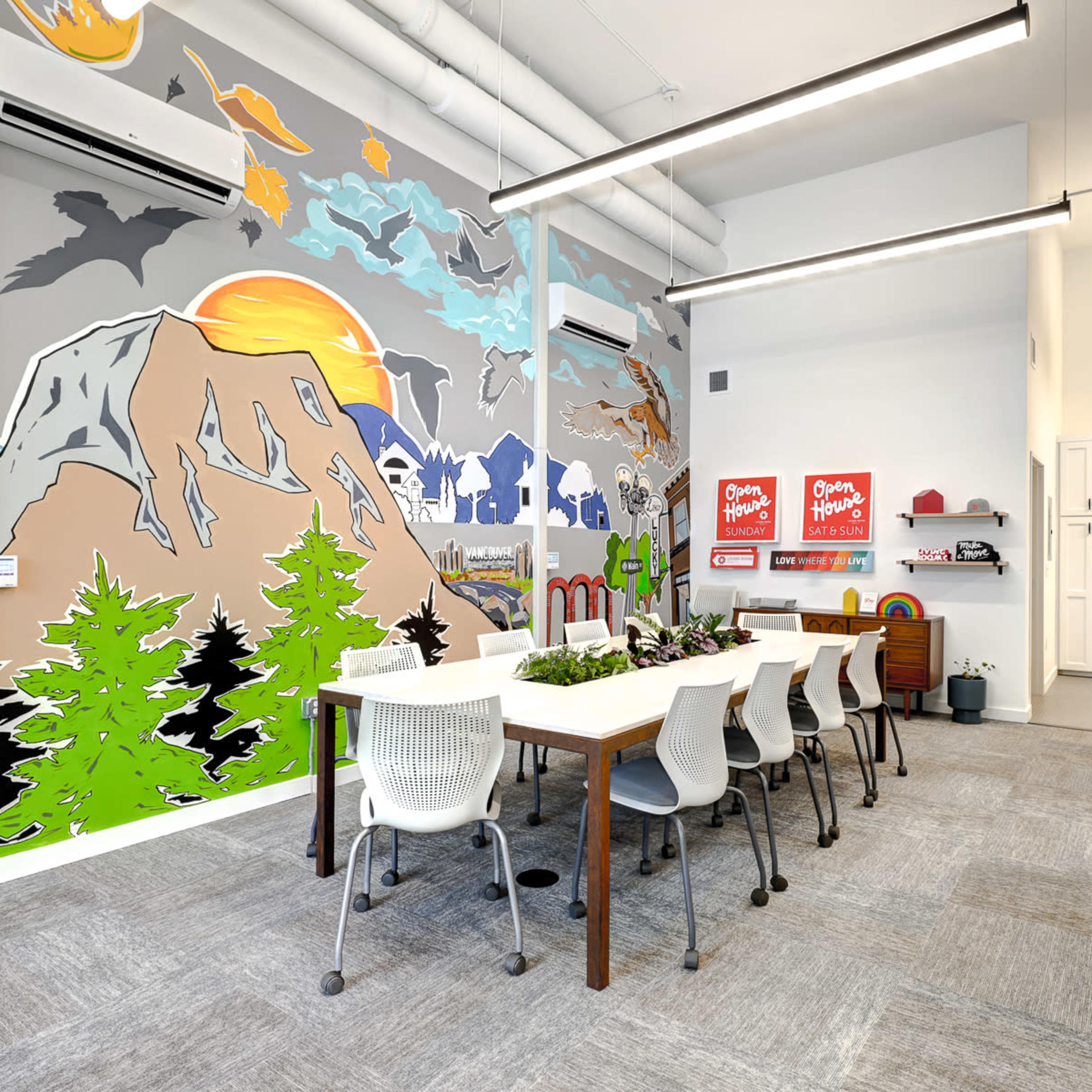 A bright meeting room features a large table surrounded by white chairs, with colorful mural artwork depicting mountains, trees, and wildlife on one wall.