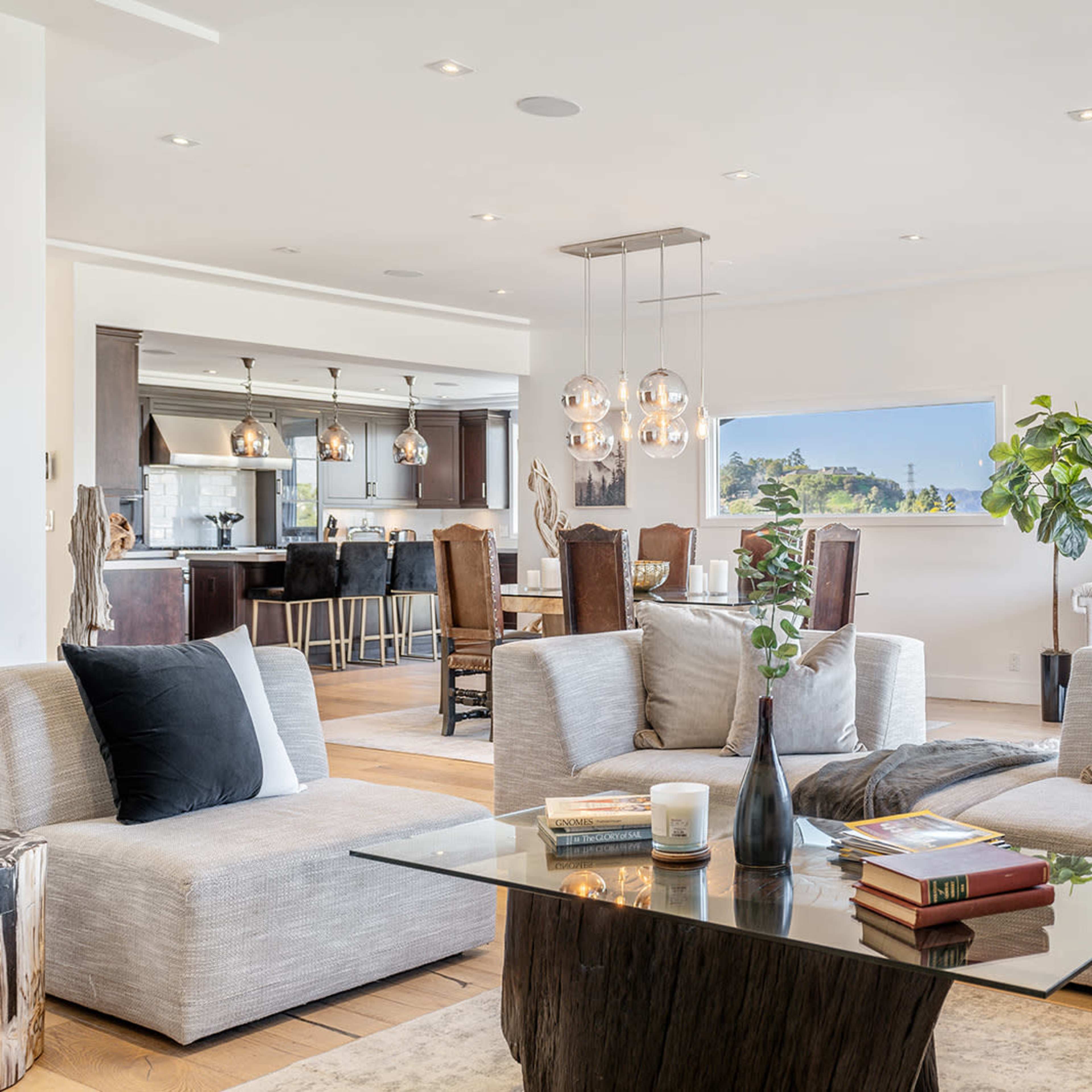 A modern living room features a spacious sectional sofa, a coffee table with books and decor, and an open view into a kitchen area with dark cabinetry and dining seating.