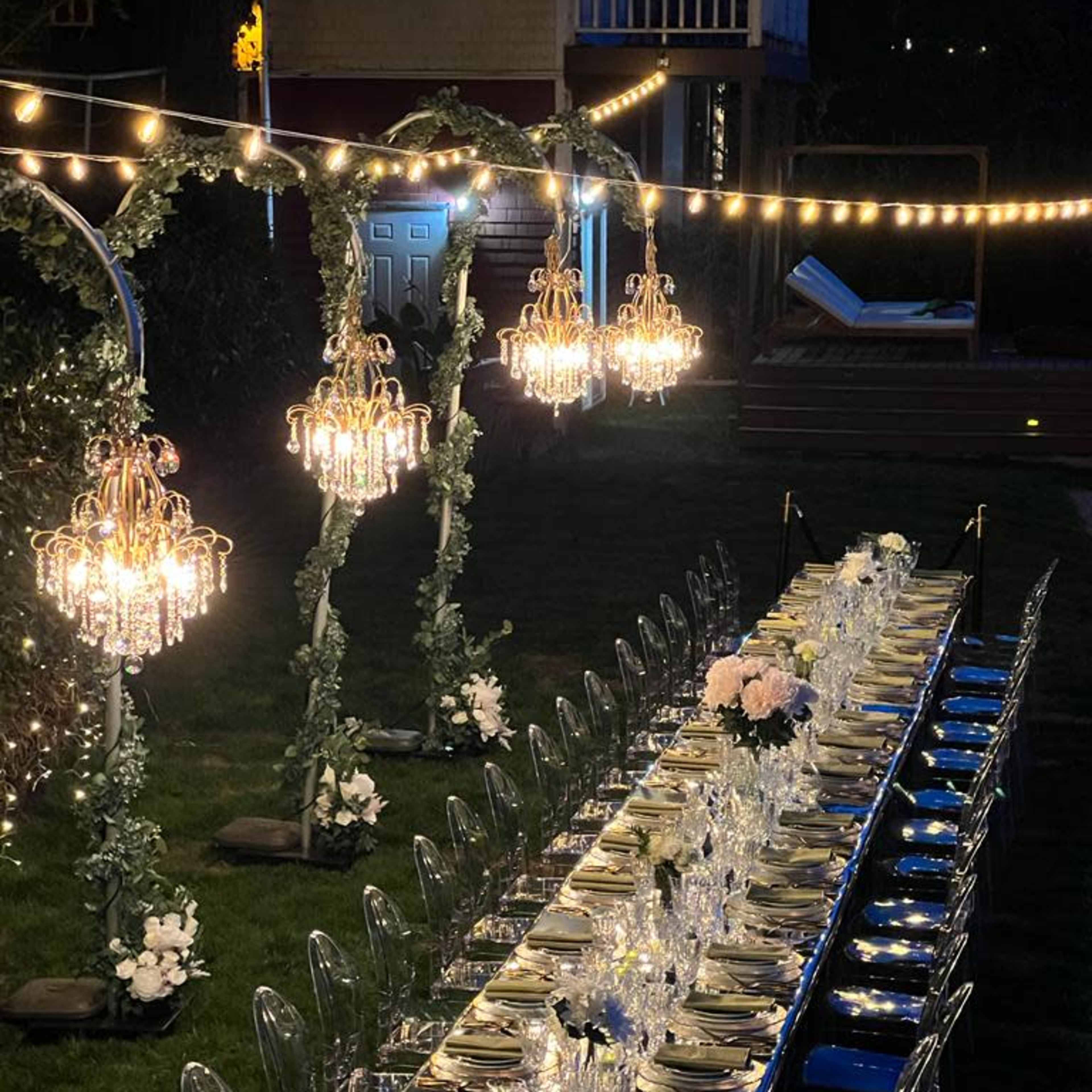 A long, elegantly set dinner table with crystal chandeliers and string lights is arranged outdoors, surrounded by greenery.