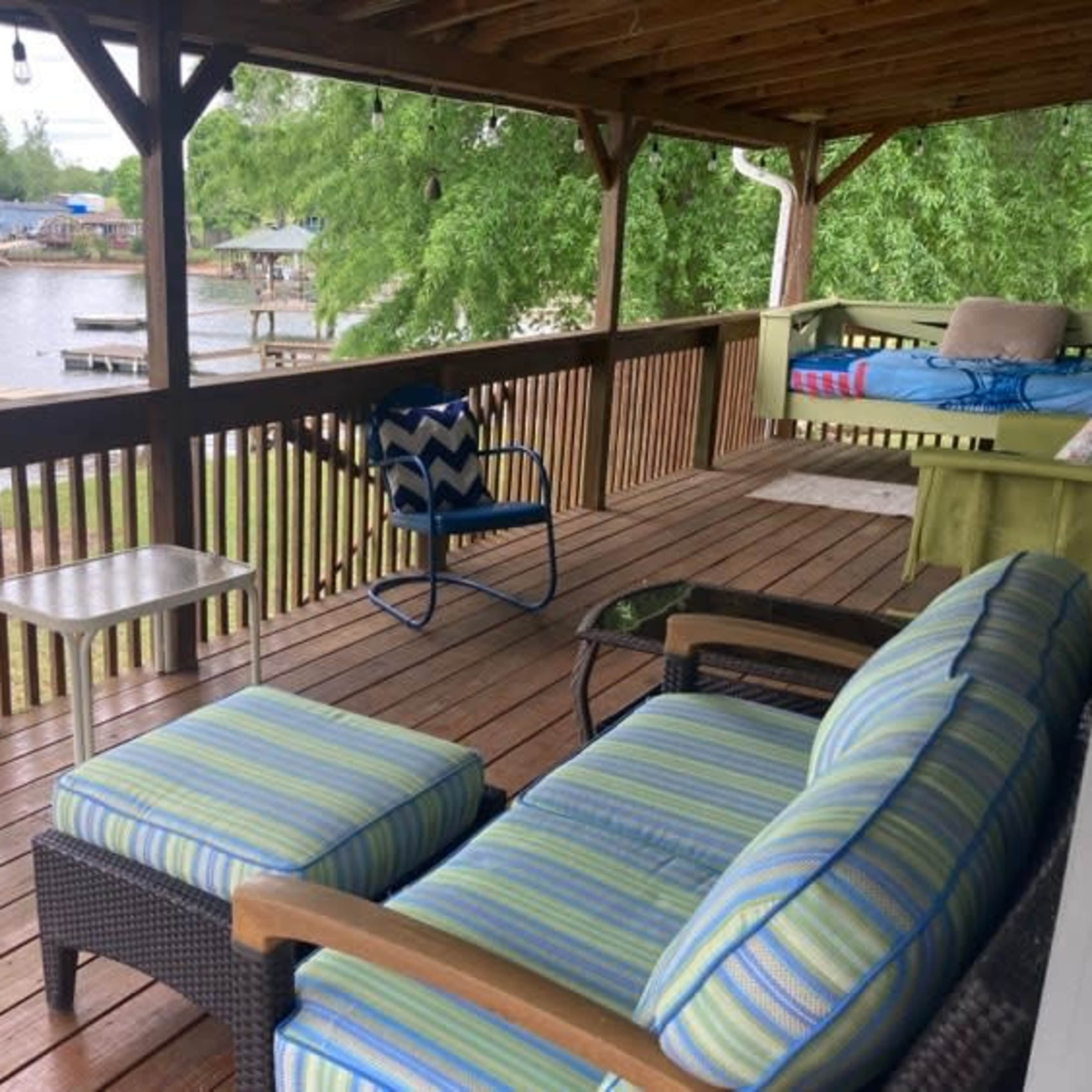 A wooden porch with a seating area that includes a striped sofa and a chair, overlooking a calm body of water with docks in the background.