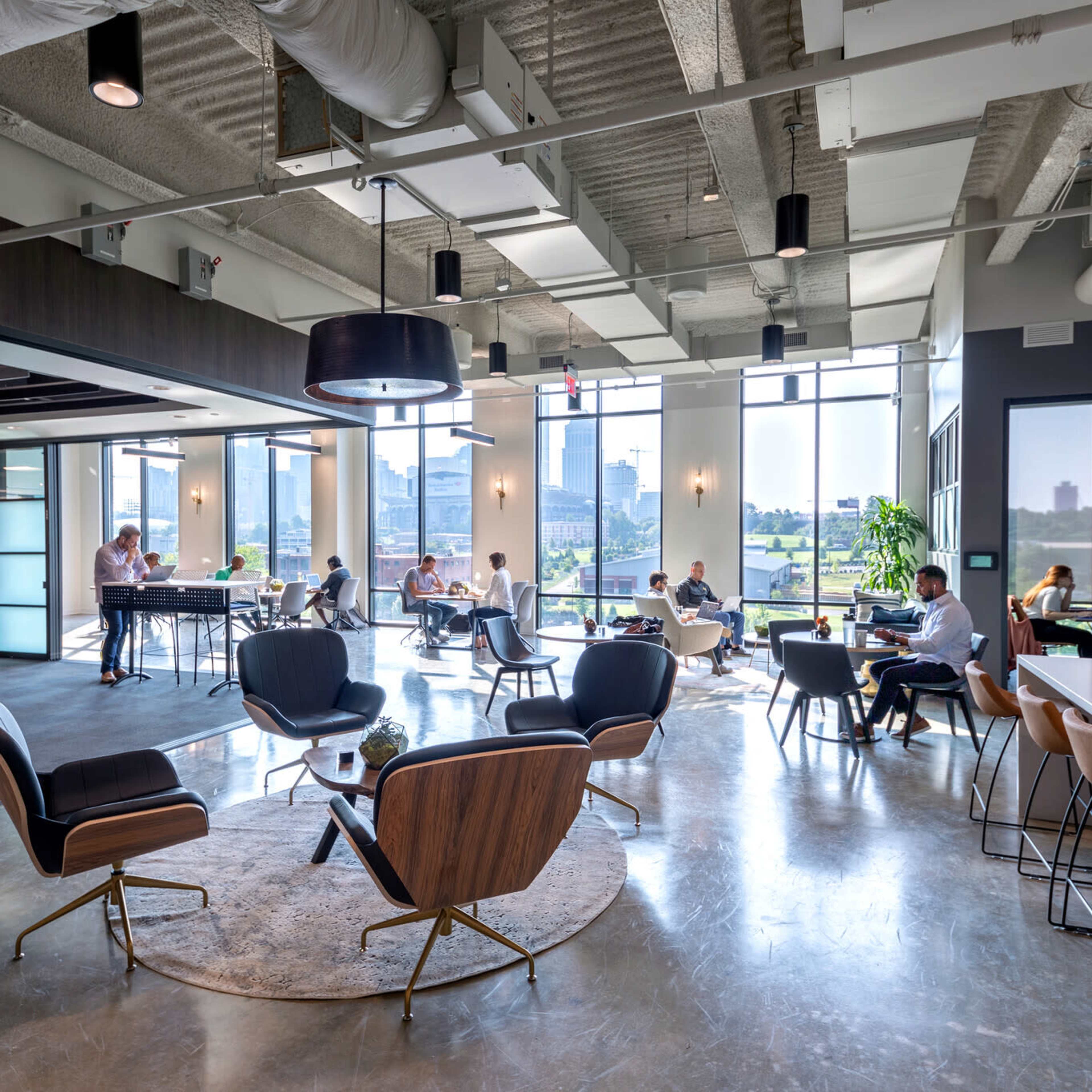 A modern office space with several people working at tables and sitting in lounge chairs, large windows allowing natural light to fill the room.