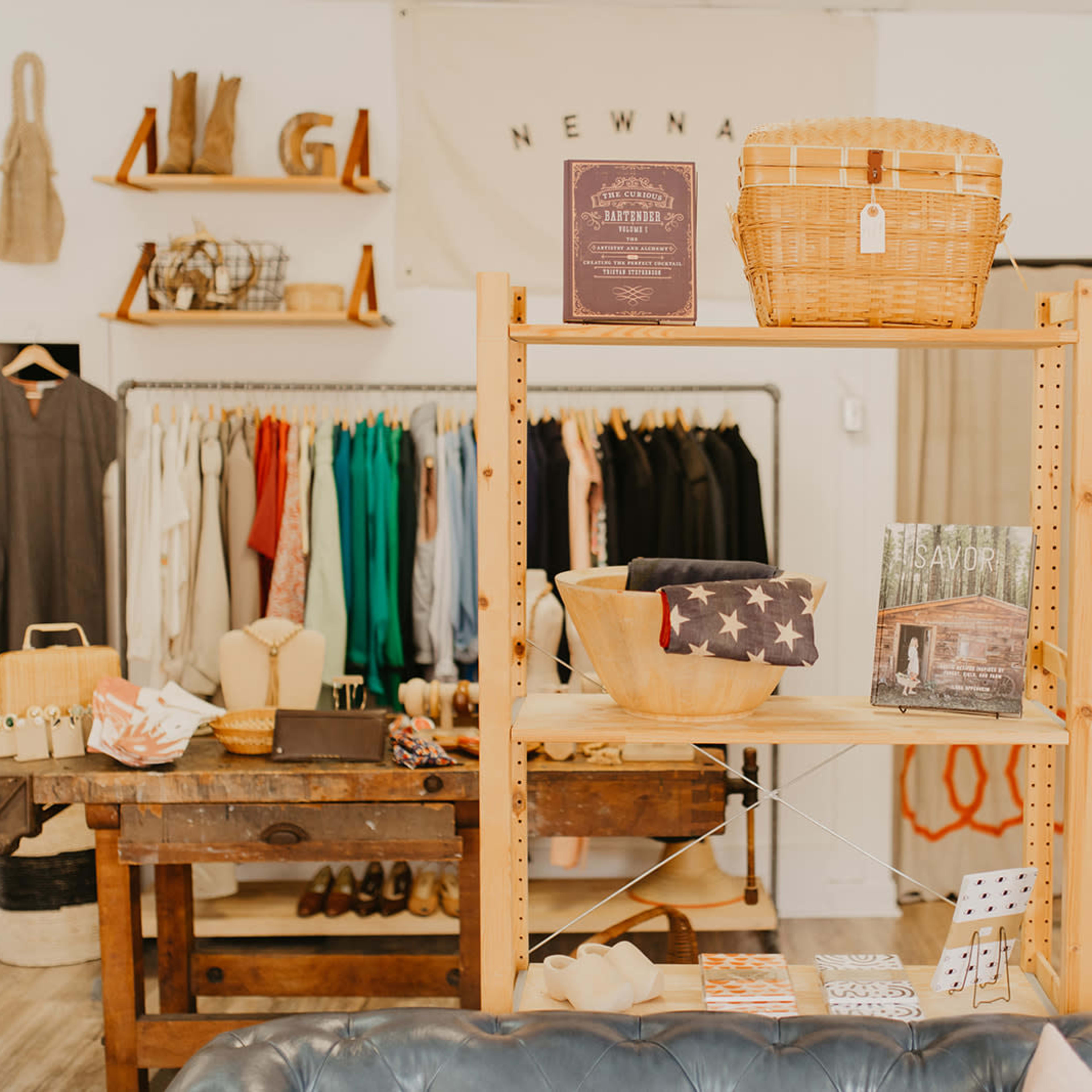 A wooden shelf displays various items, including a wicker basket, bowls, books, and accessories, in a retail space with clothing on hangers in the background.