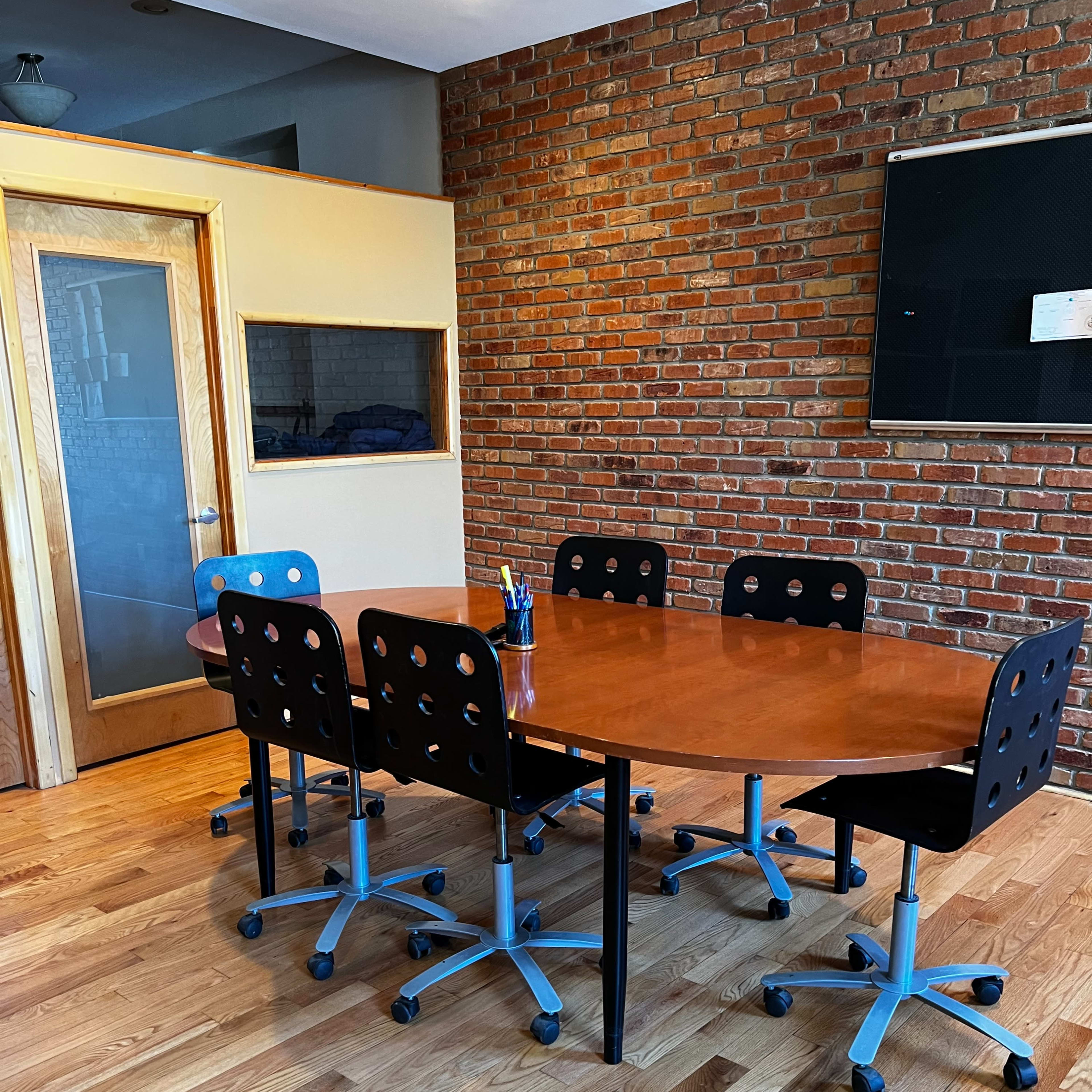 A conference room features a wooden table surrounded by six black office chairs on casters, with a brick wall in the background and a glass-panel door.