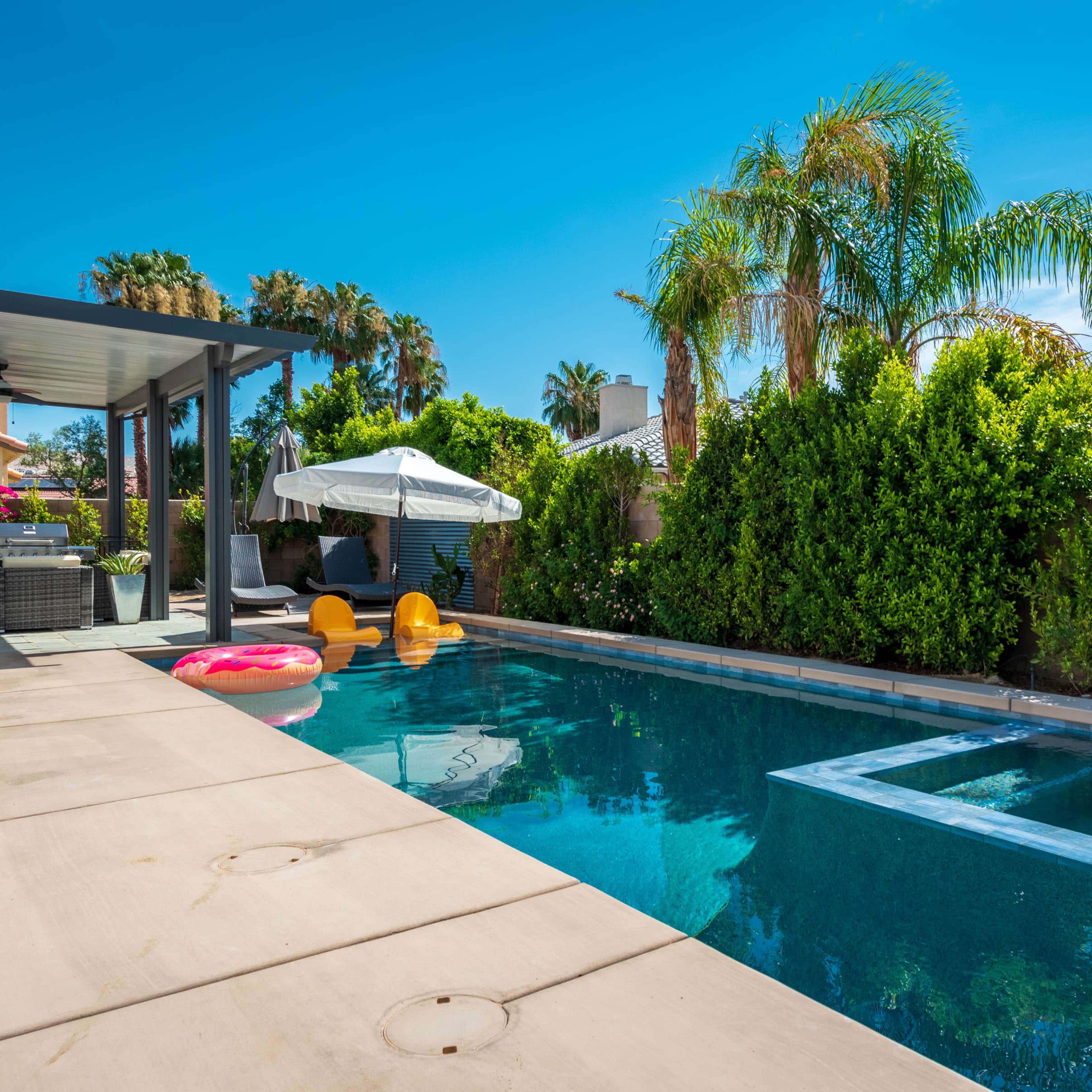 A backyard pool surrounded by palm trees, shrubs, and outdoor furniture, with inflatable floats on the water.