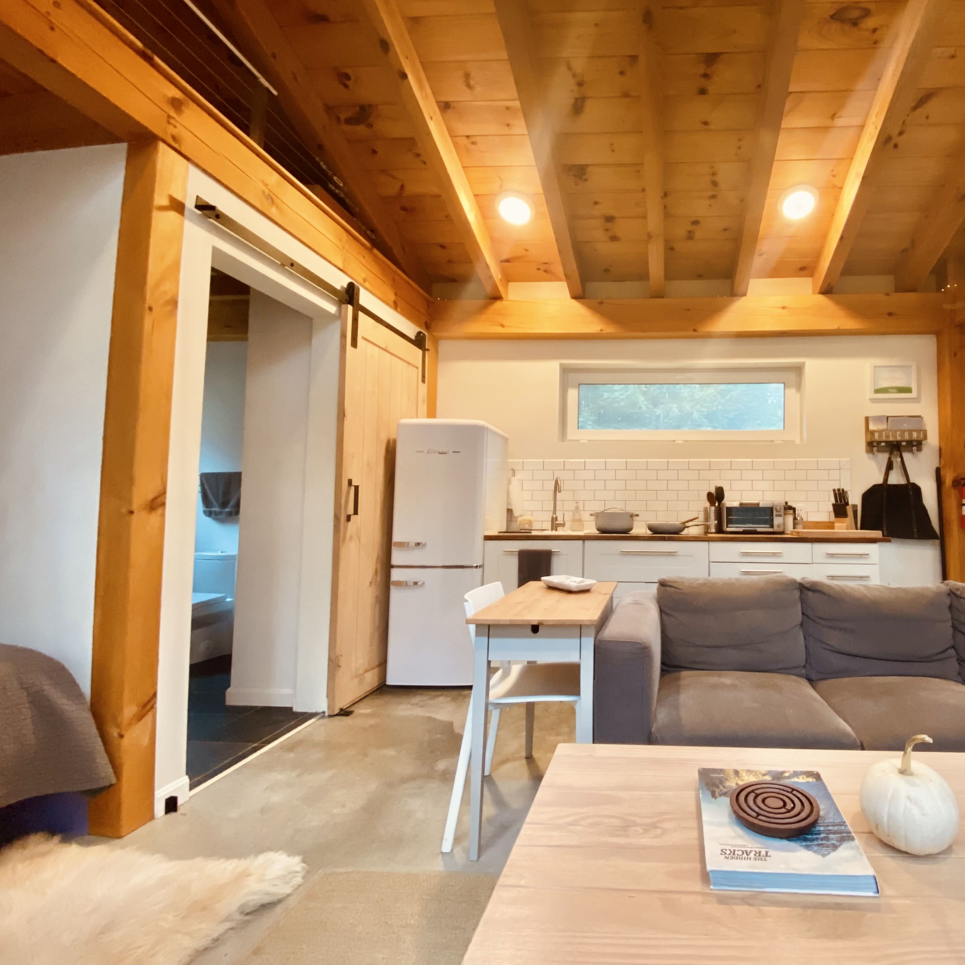 A cozy interior of a cabin with a wooden ceiling, a gray sectional sofa, a small dining table, and a kitchen area featuring a white refrigerator and a tiled backsplash.