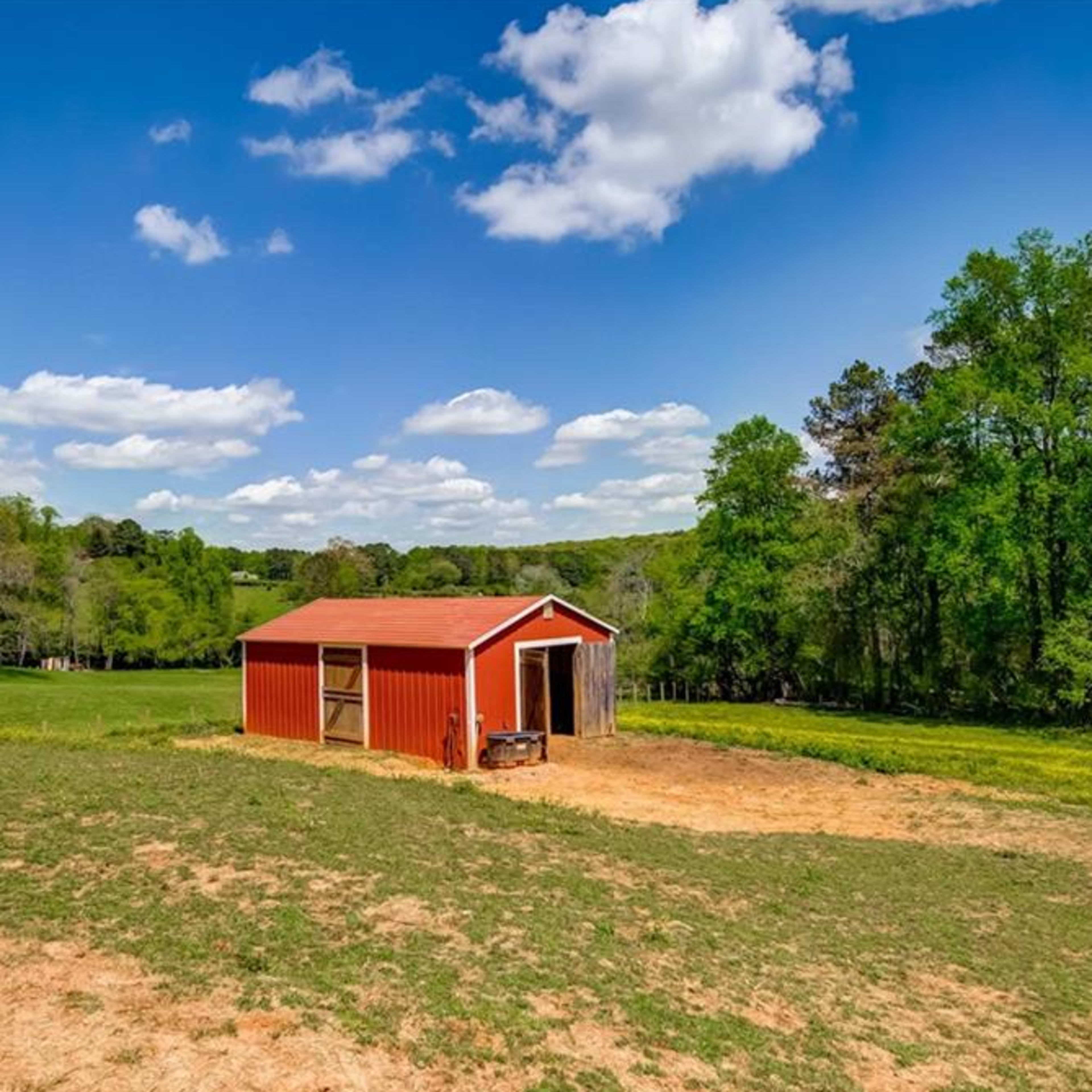 A red barn is situated in a grassy field surrounded by trees under a blue sky with scattered clouds.