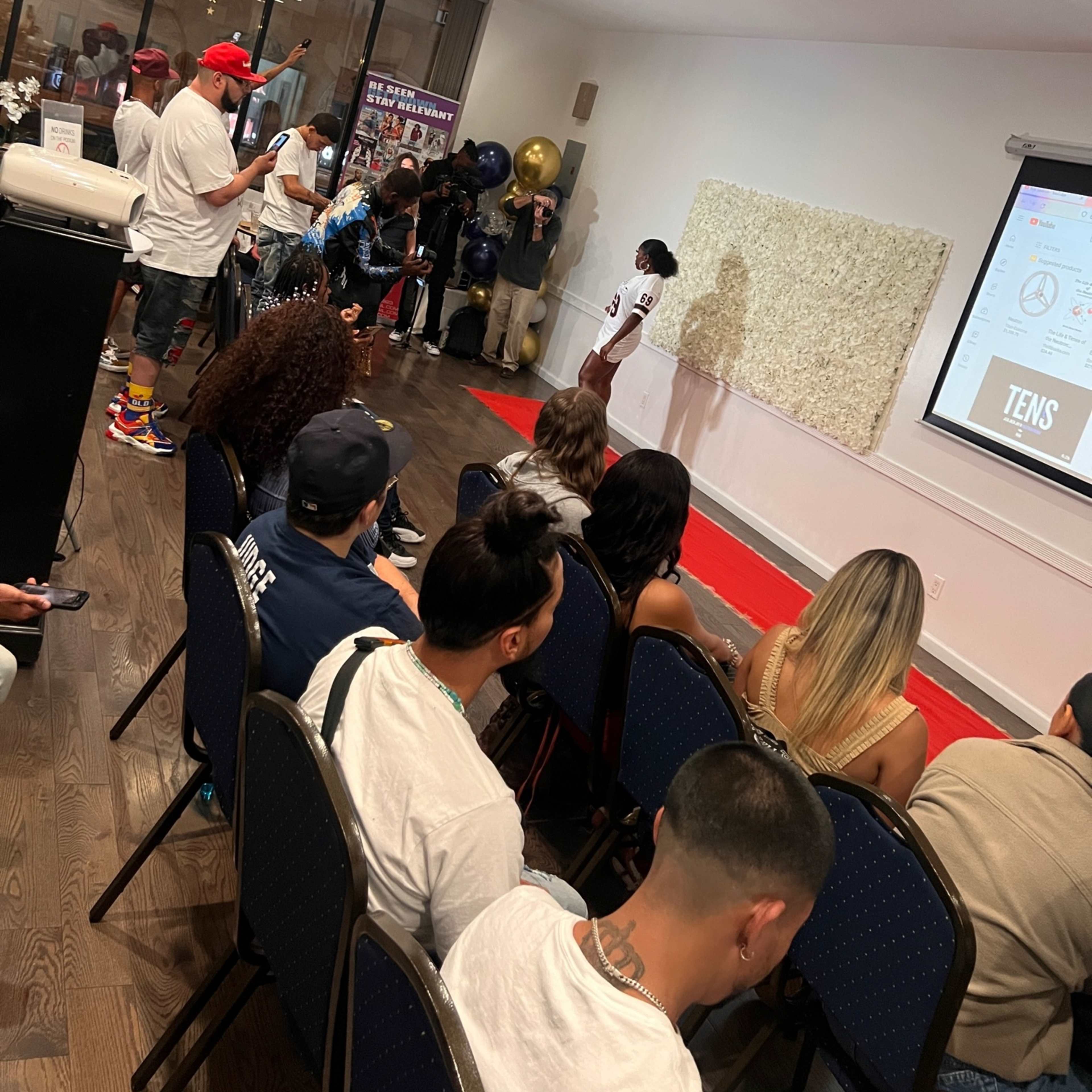 A group of people watches a presentation in a well-lit room with a red carpet and a projector screen displaying information.