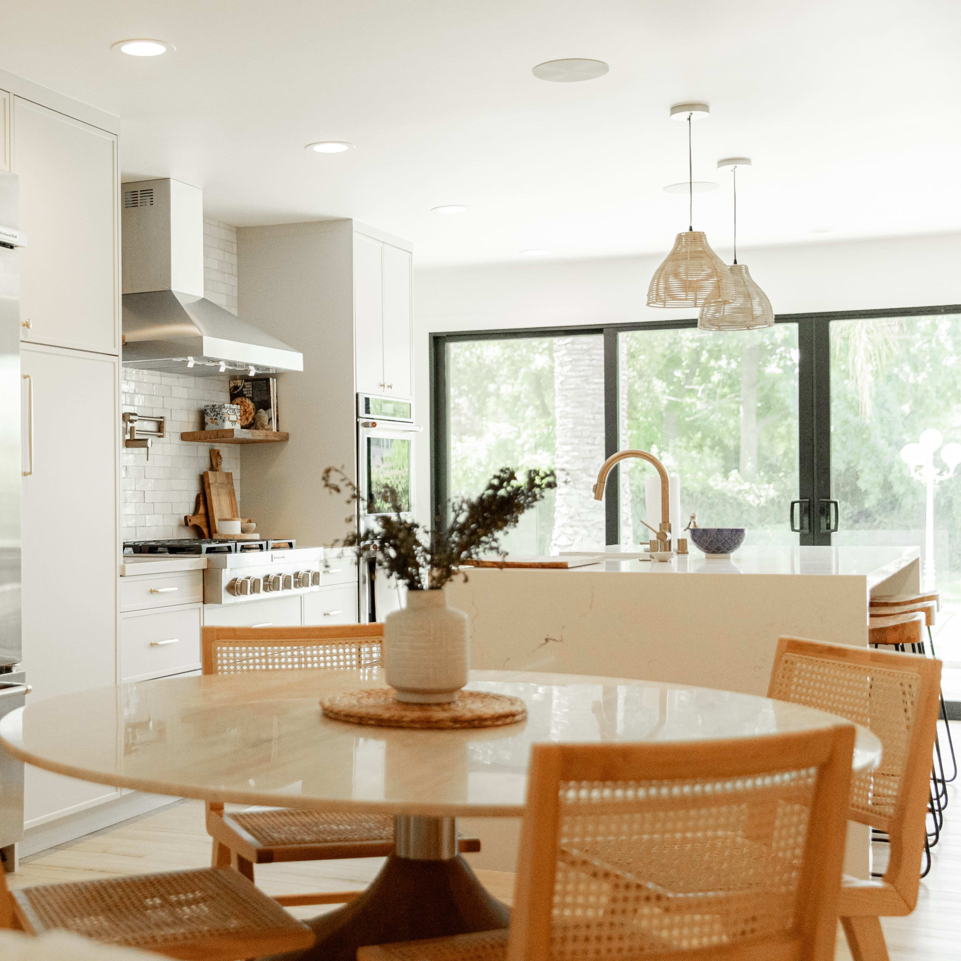 A modern kitchen features stainless steel appliances, a large island with seating, and a dining table surrounded by woven chairs.