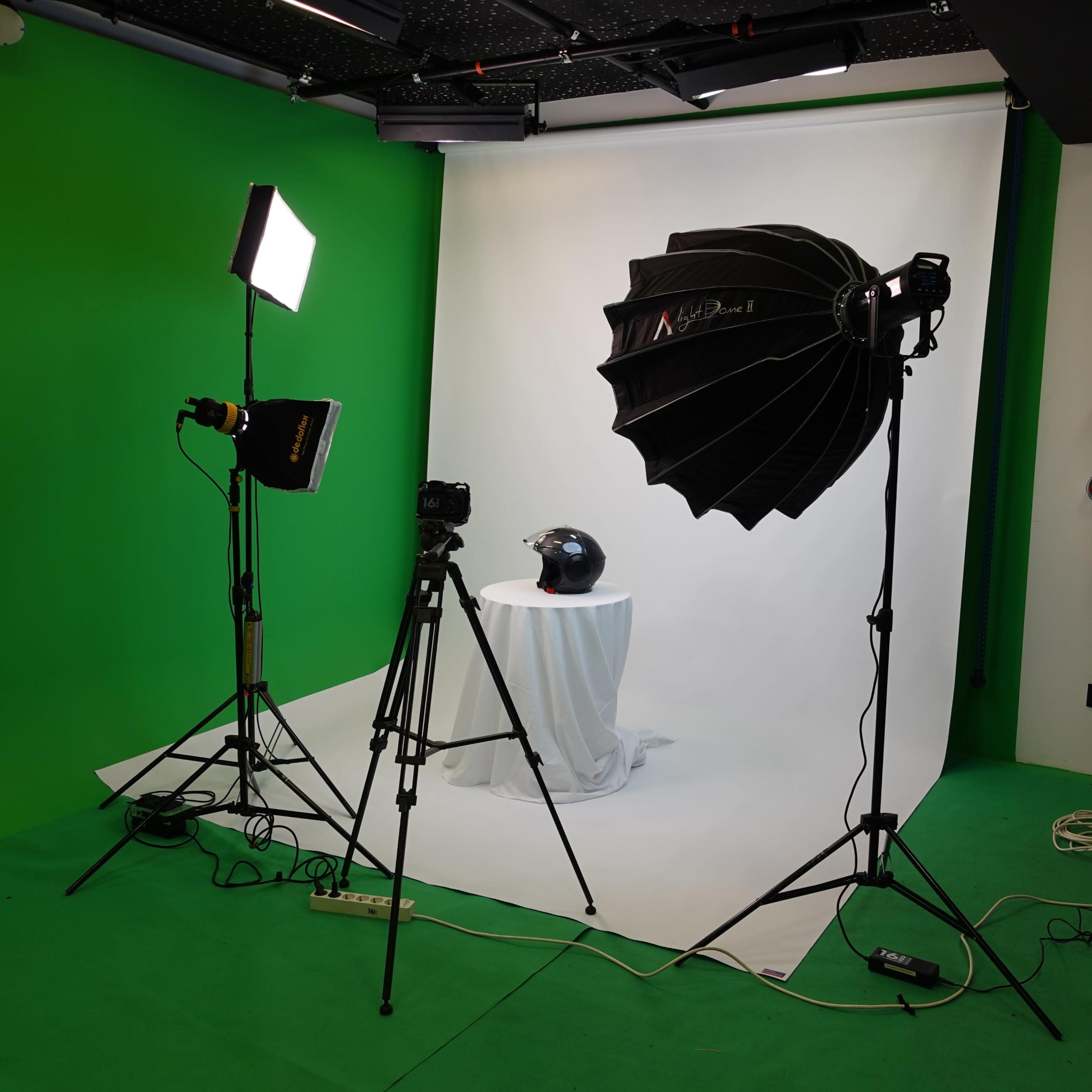 A photography studio setup with a green backdrop, a softbox light, a camera on a tripod, and a helmet placed on a pedestal.