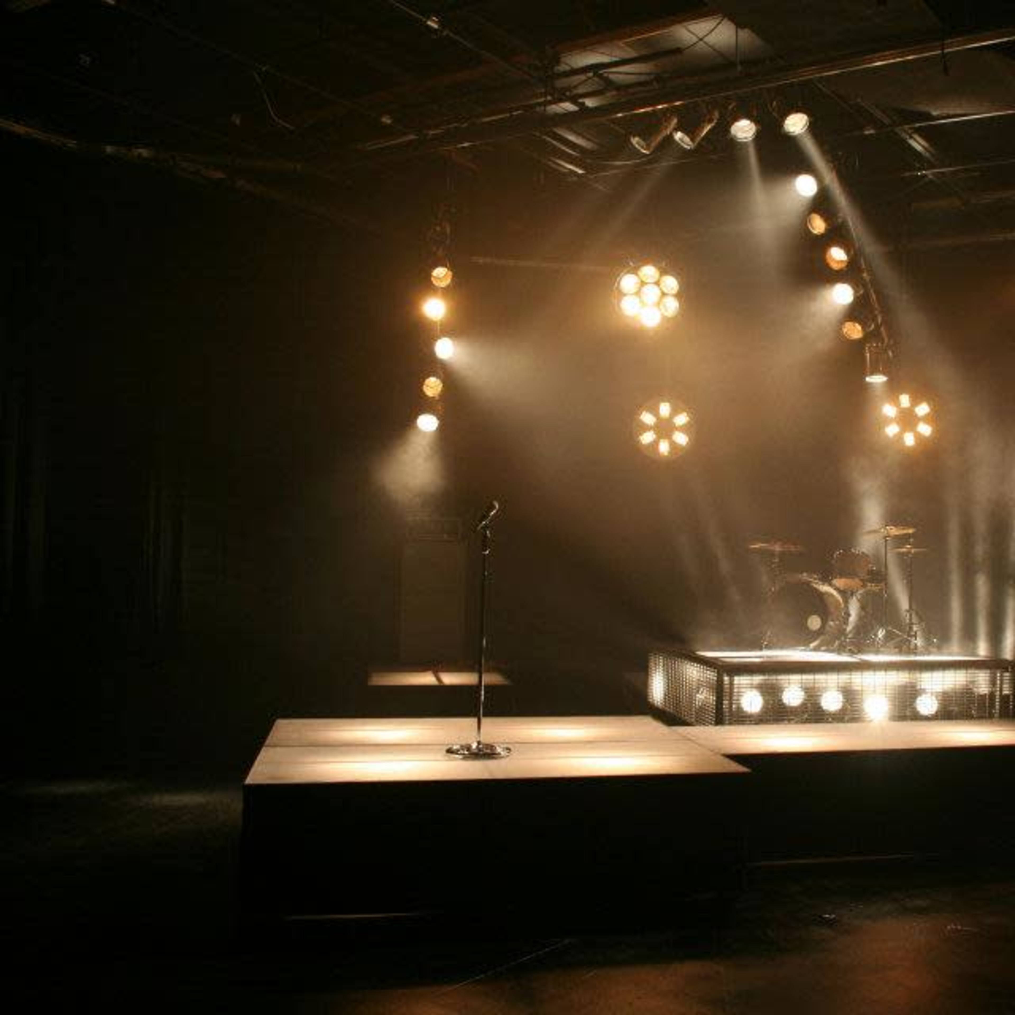 A dimly lit stage is set with spotlights, a microphone stand, and a drum kit surrounded by large amplifiers.