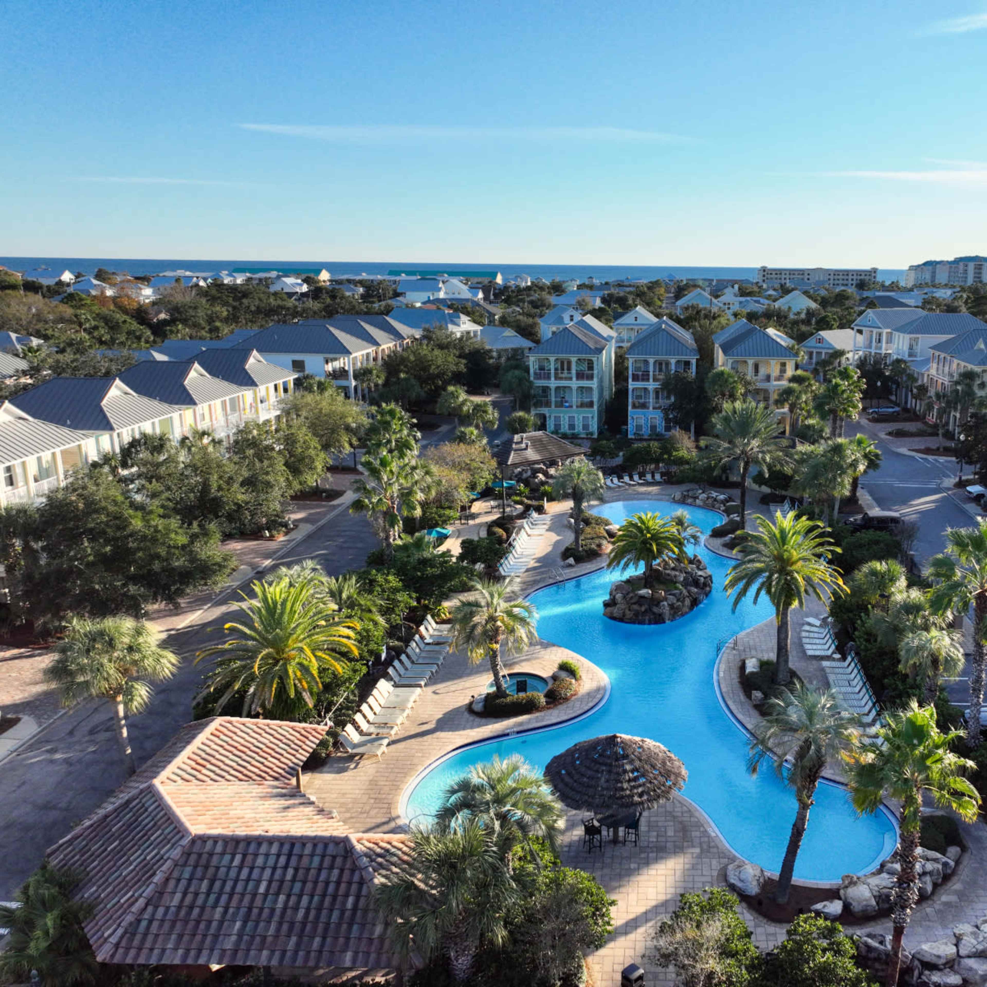A bird's-eye view of a resort-style pool surrounded by palm trees and residential buildings, with the ocean visible in the background.