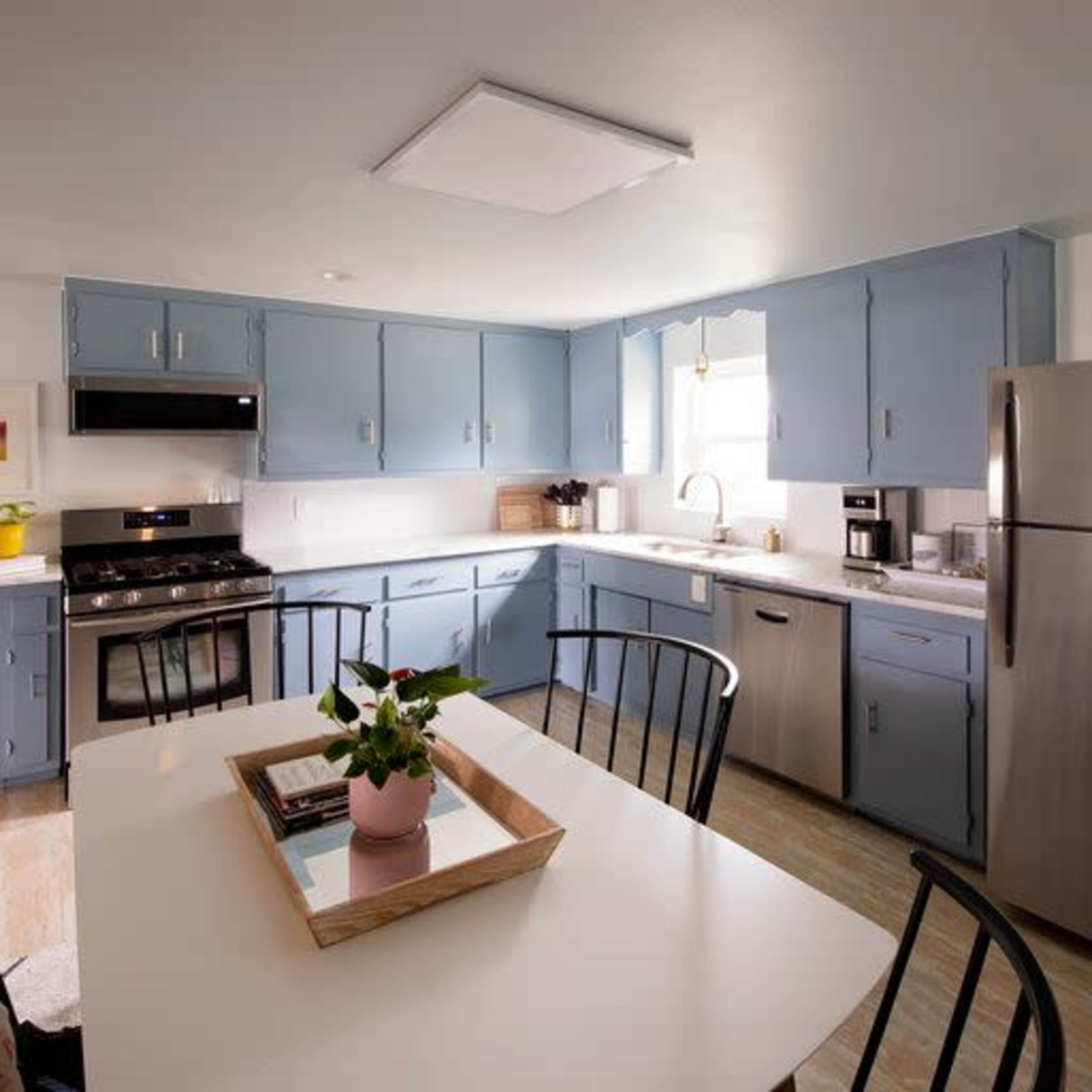 A bright kitchen features blue cabinets, stainless steel appliances, and a dining table with a small plant centerpiece.