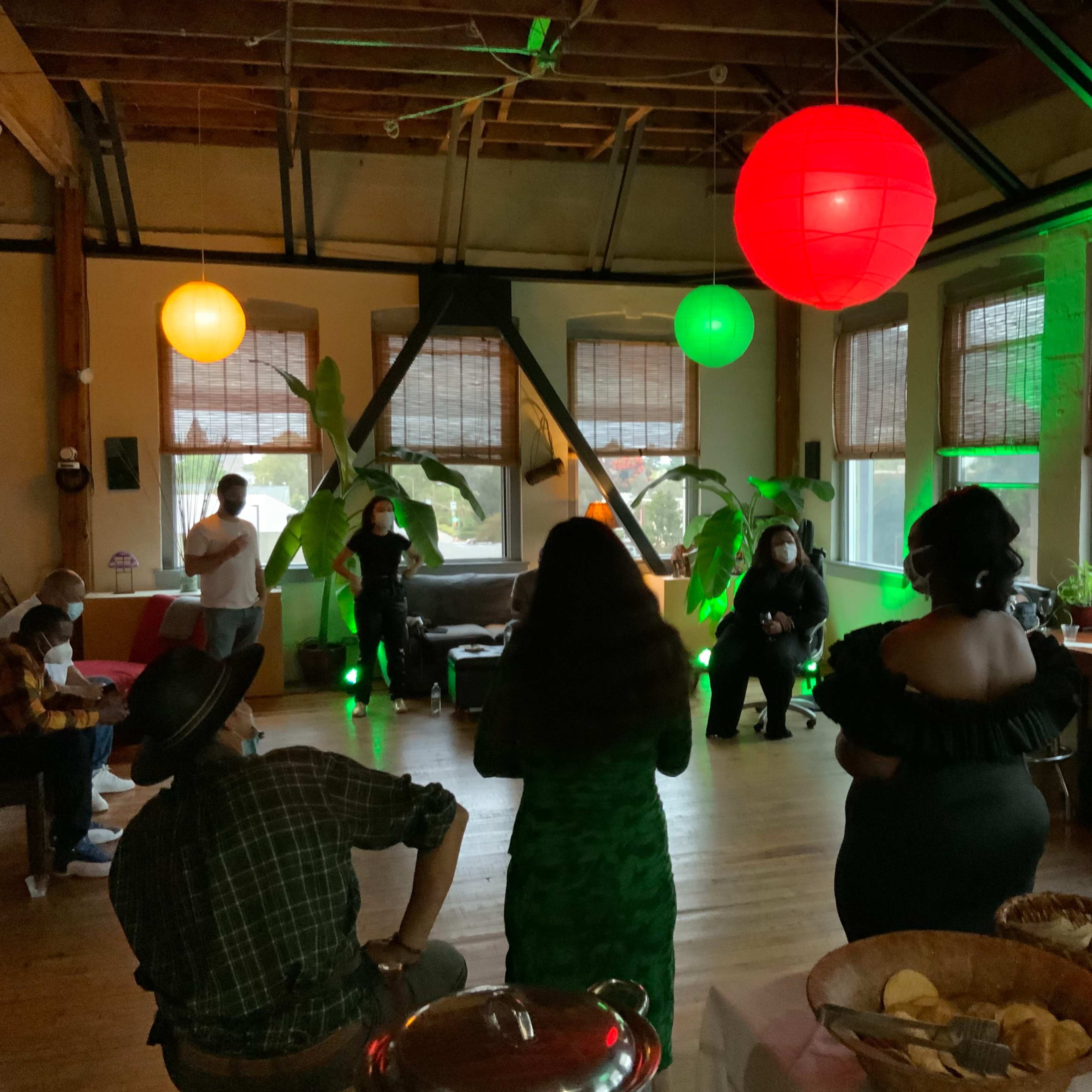 A group of people stands in a spacious room with wooden floors and large windows, while colorful lanterns hang overhead and green lights illuminate the space.