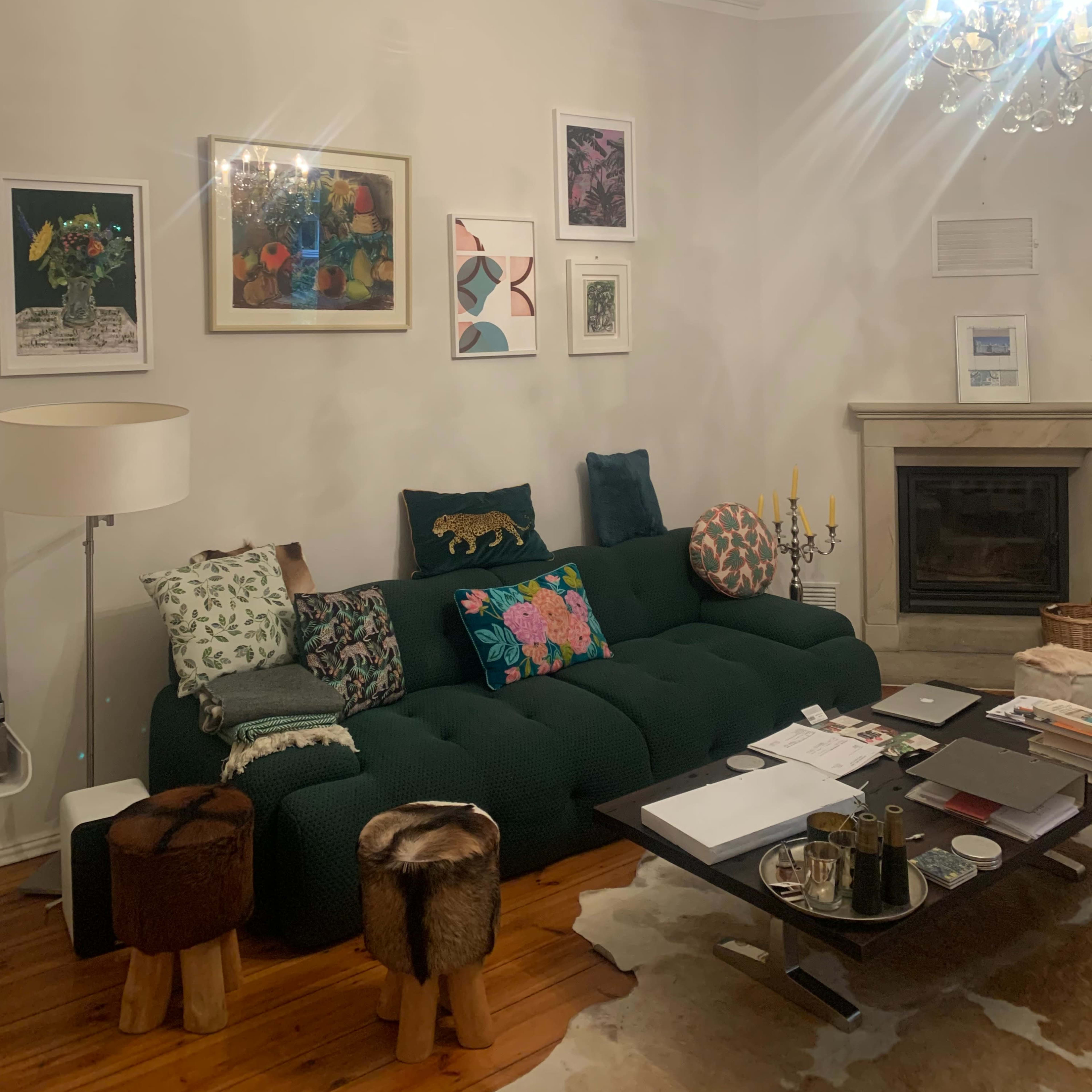 A cozy living room features a green sofa adorned with colorful cushions, a coffee table cluttered with books and decorative items, and various artworks displayed on the walls.