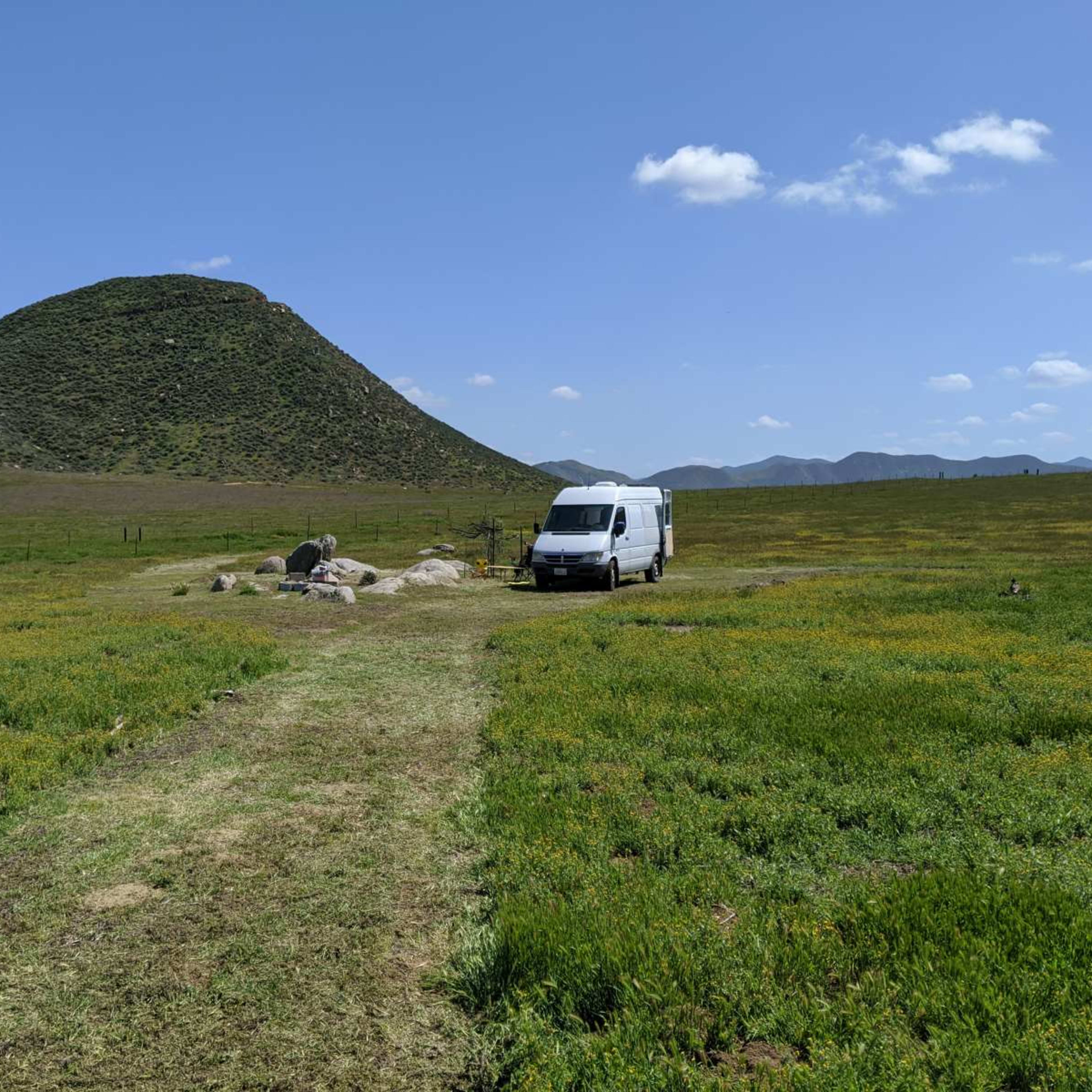 A white van is parked on a dirt path surrounded by green fields and a large hill in the background under a blue sky.