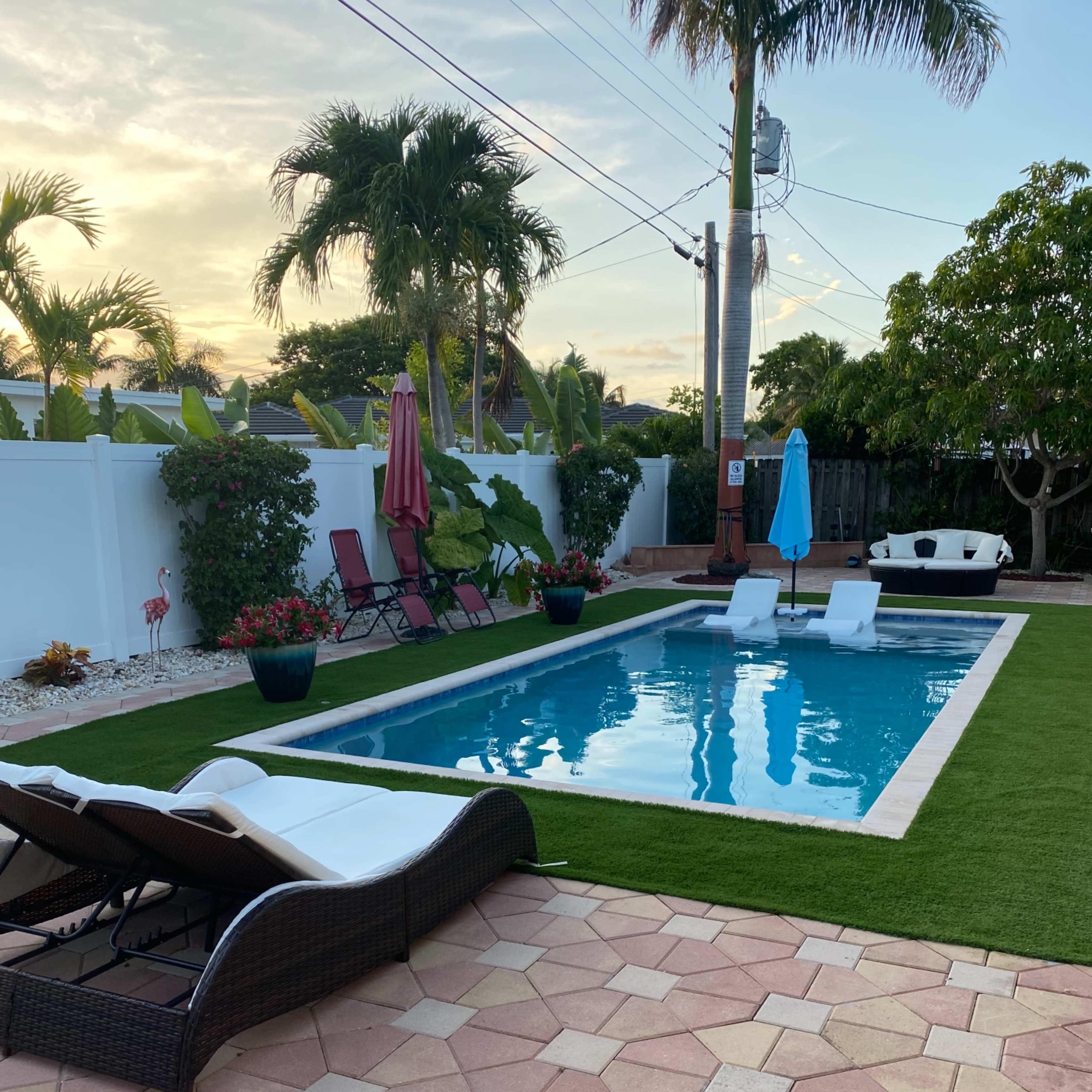 A backyard pool area with lounge chairs, a landscaped yard, and palm trees, surrounded by a white fence.