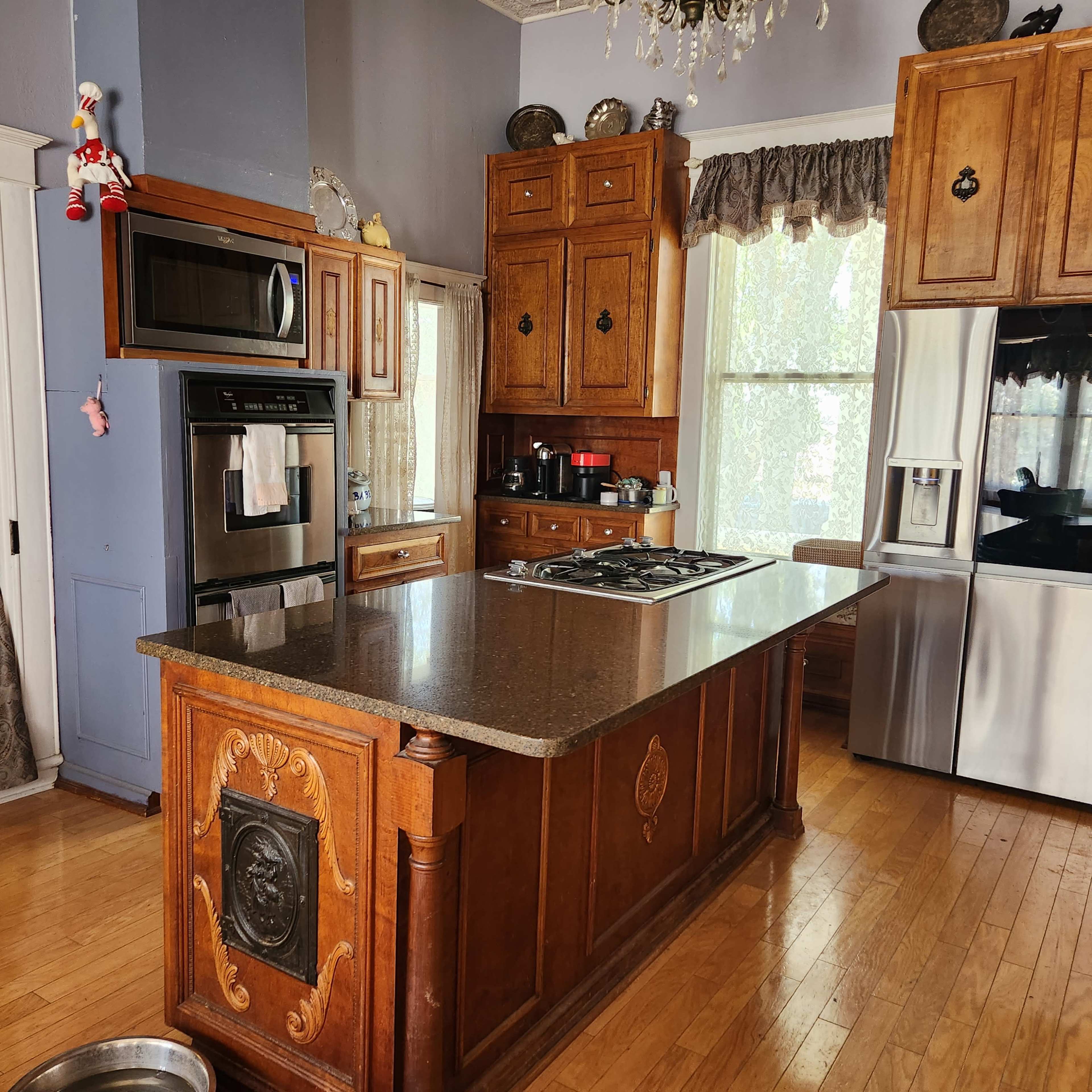 A spacious kitchen with wooden cabinetry, a central island with a cooktop, stainless steel appliances, and a staircase leading to an upper floor.