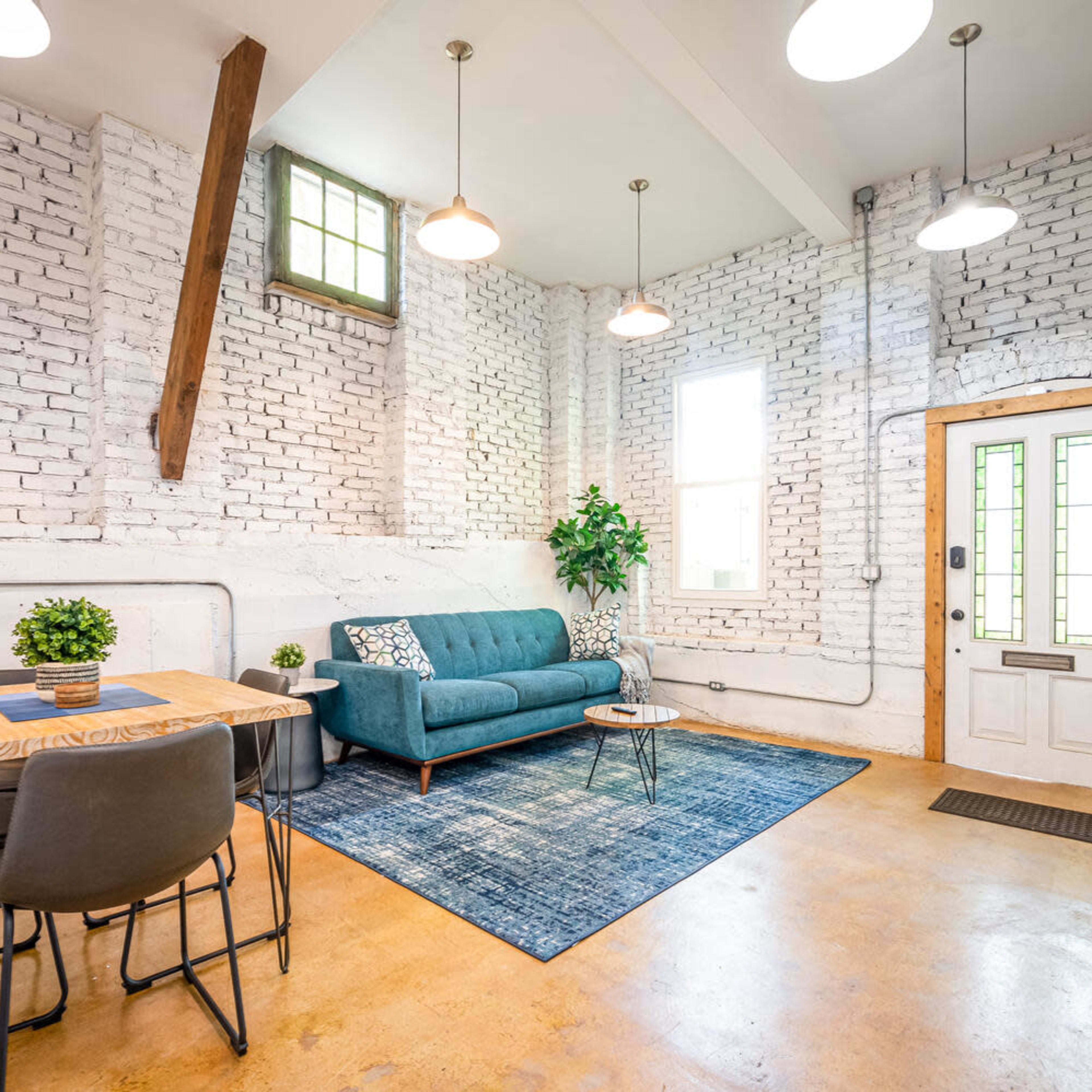 A modern living space featuring a teal couch, a wooden dining table with chairs, and a large area rug on a concrete floor, with white brick walls and large windows allowing natural light.