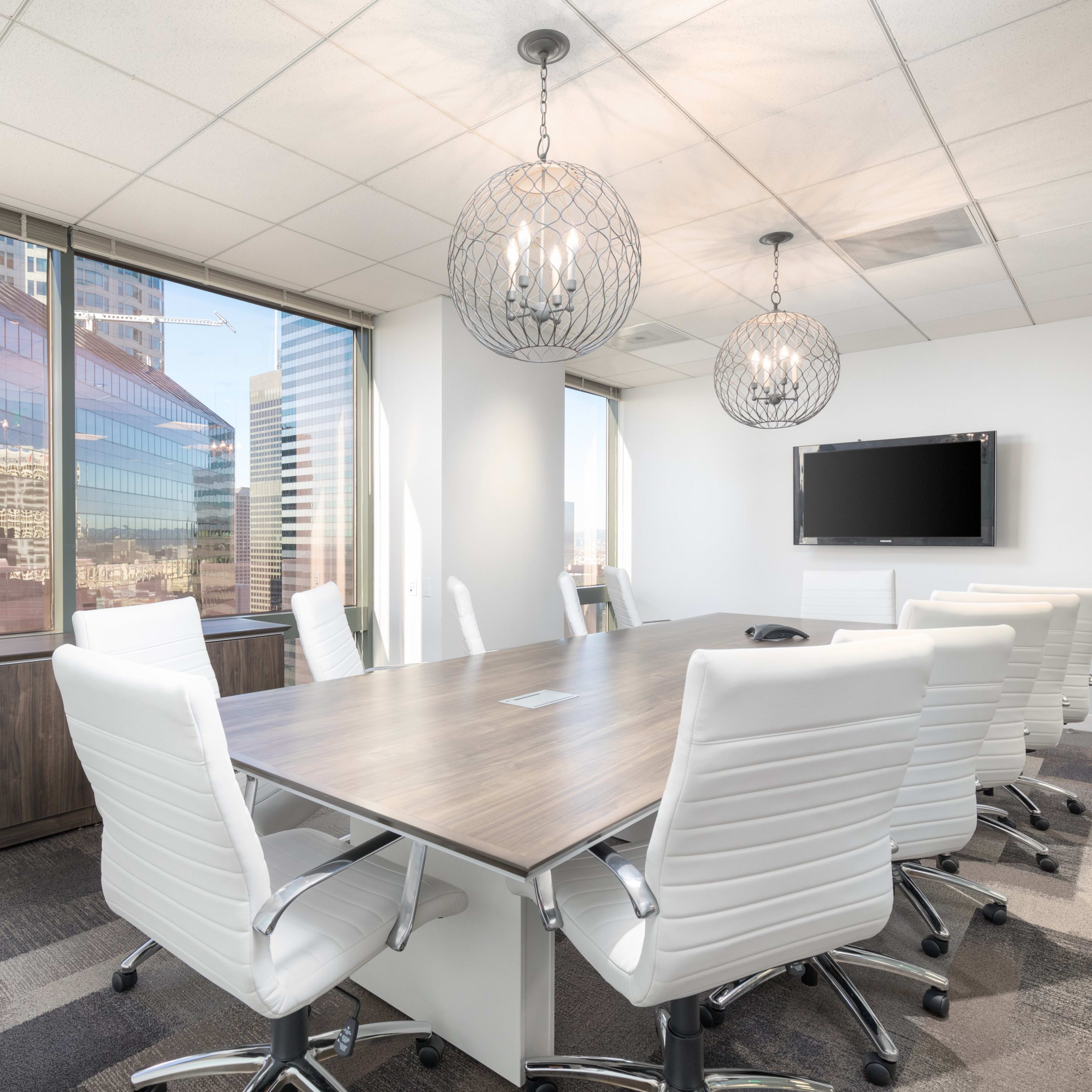 A modern conference room with a large wooden table surrounded by white chairs, large windows revealing a city view, and two pendant lights overhead.