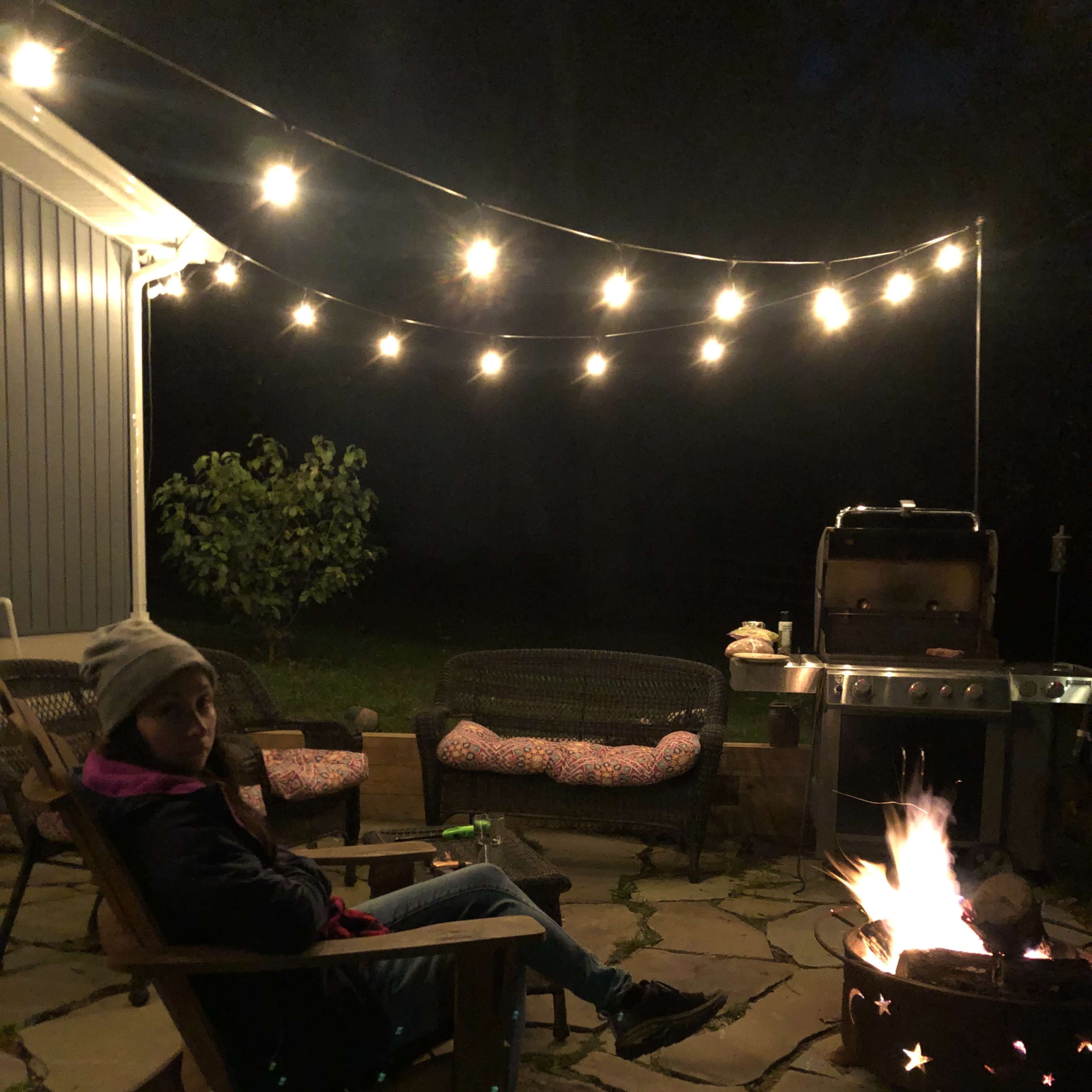 A person sits on an outdoor chair by a fire pit surrounded by string lights in a backyard at night.
