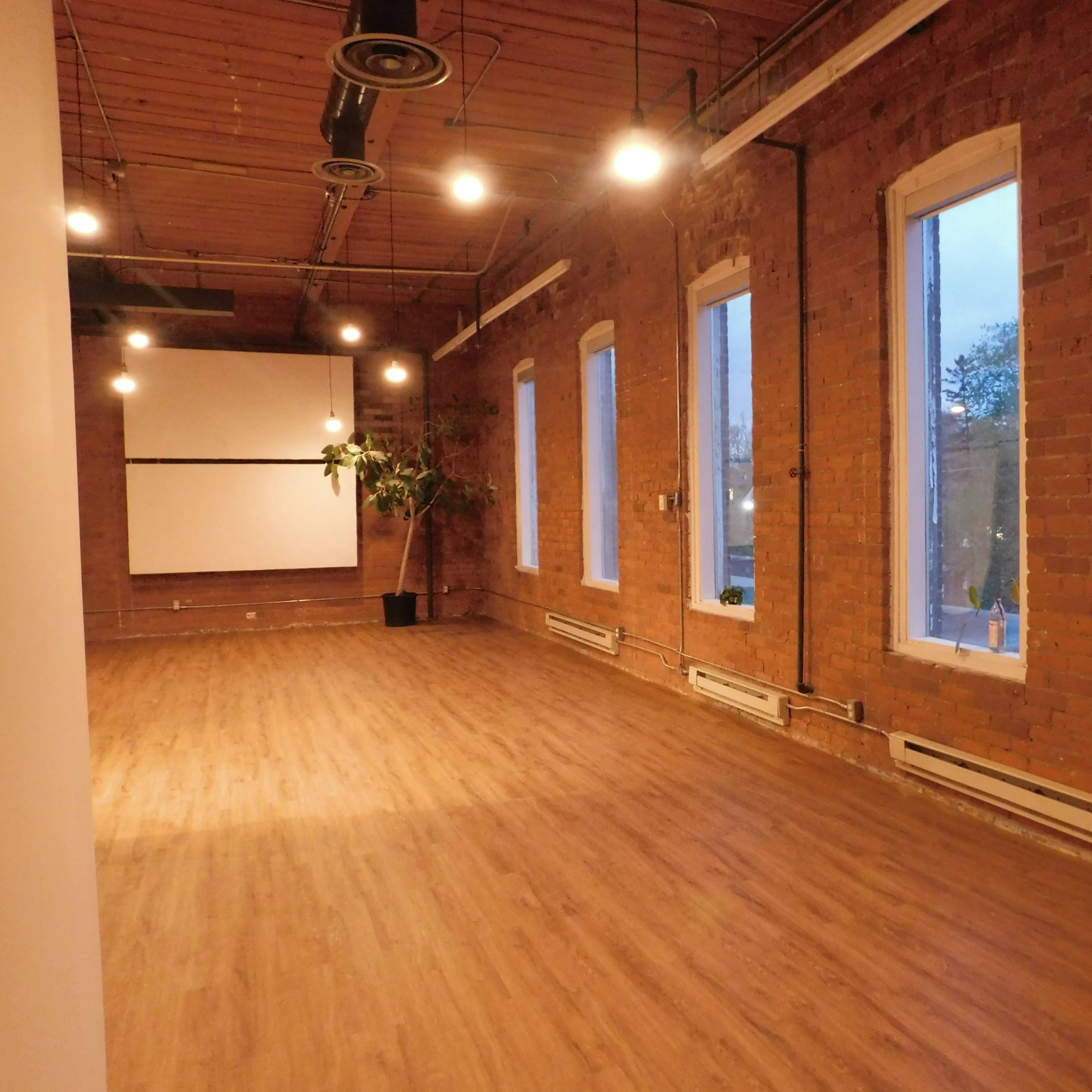 An empty room with exposed brick walls, hardwood flooring, large windows, and a projector screen mounted on the far wall.