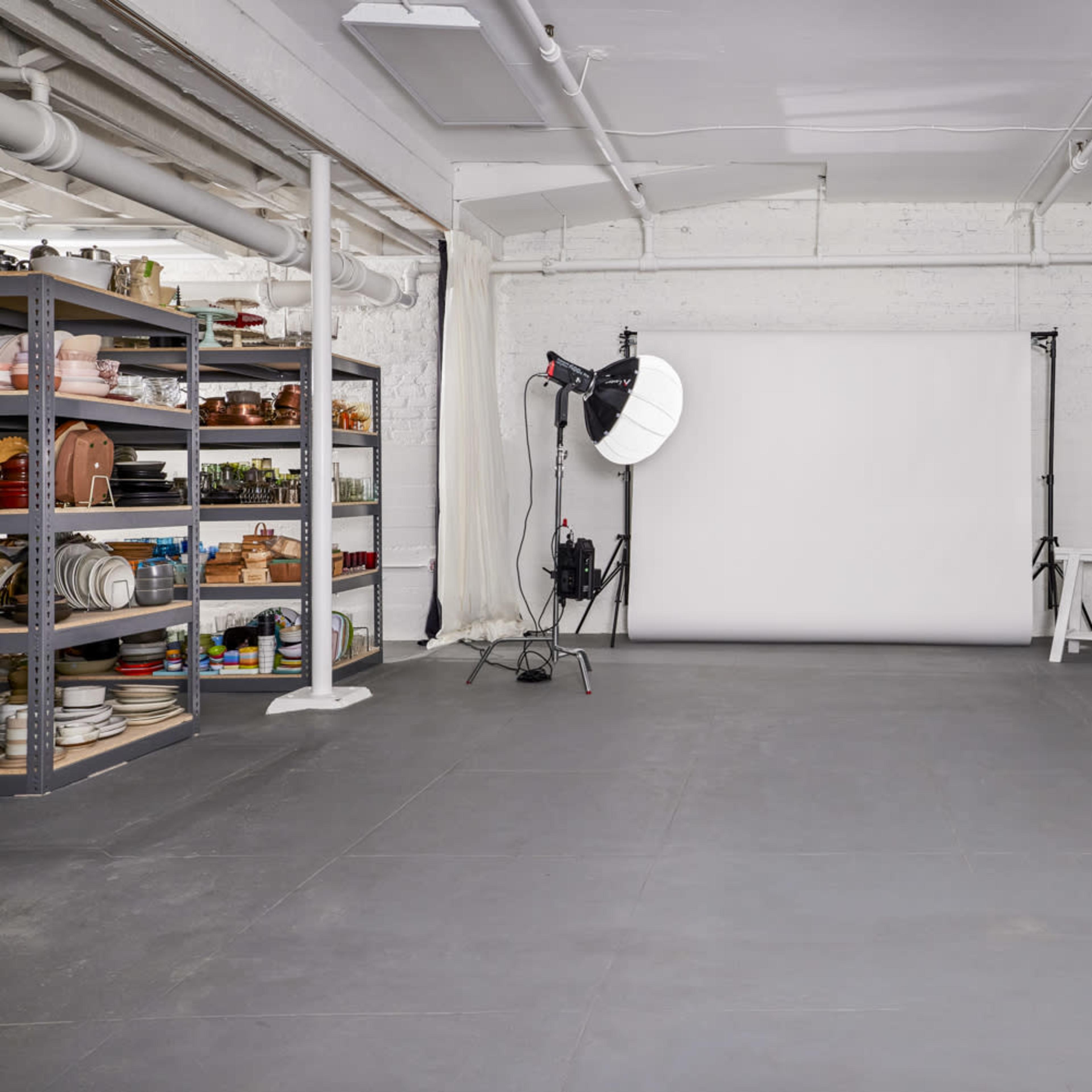 A spacious photography studio with a blank backdrop, lighting equipment, a shelving unit filled with various props, and a large mirror.