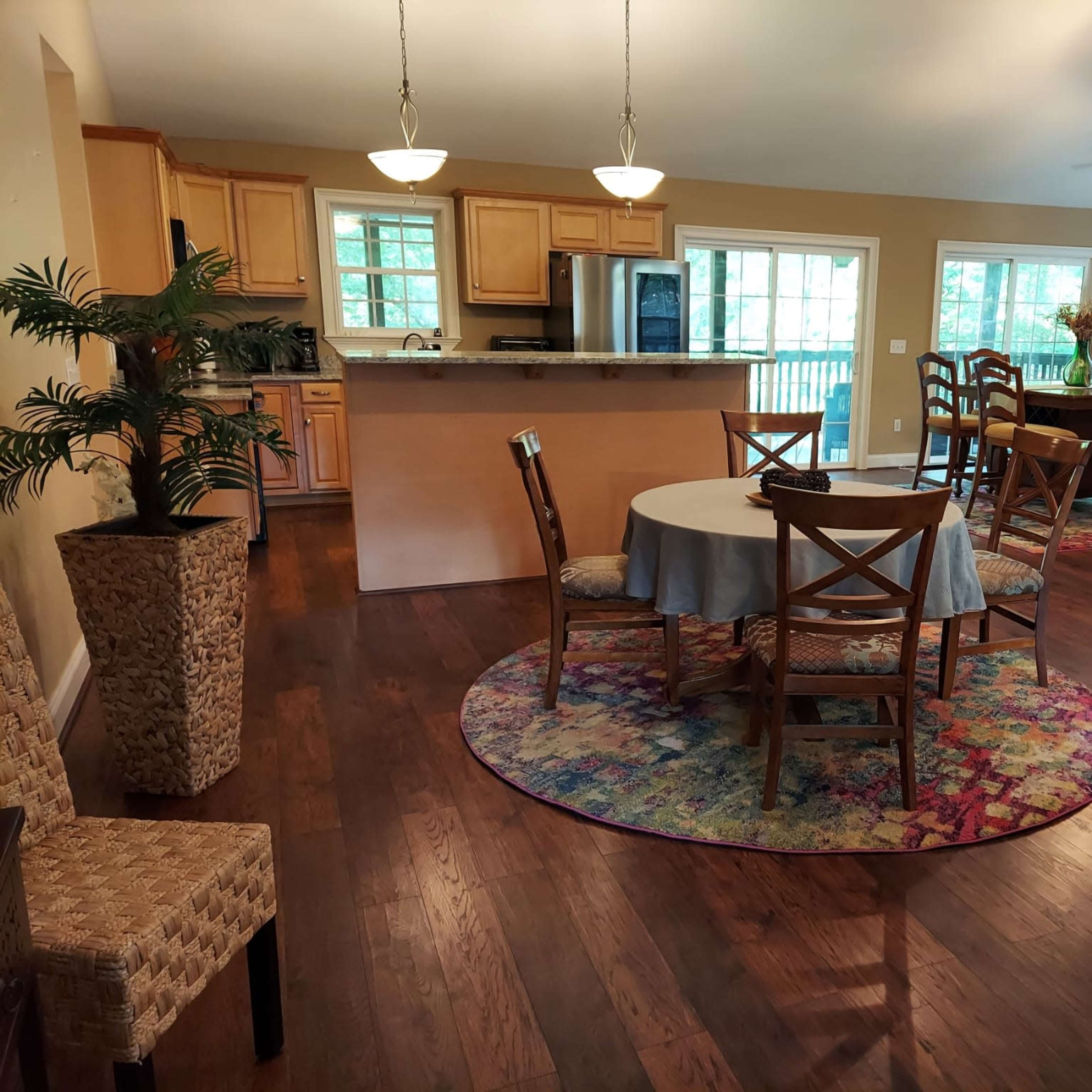 A spacious living area features a dining table surrounded by chairs, with a kitchen in the background and large windows illuminating the space.