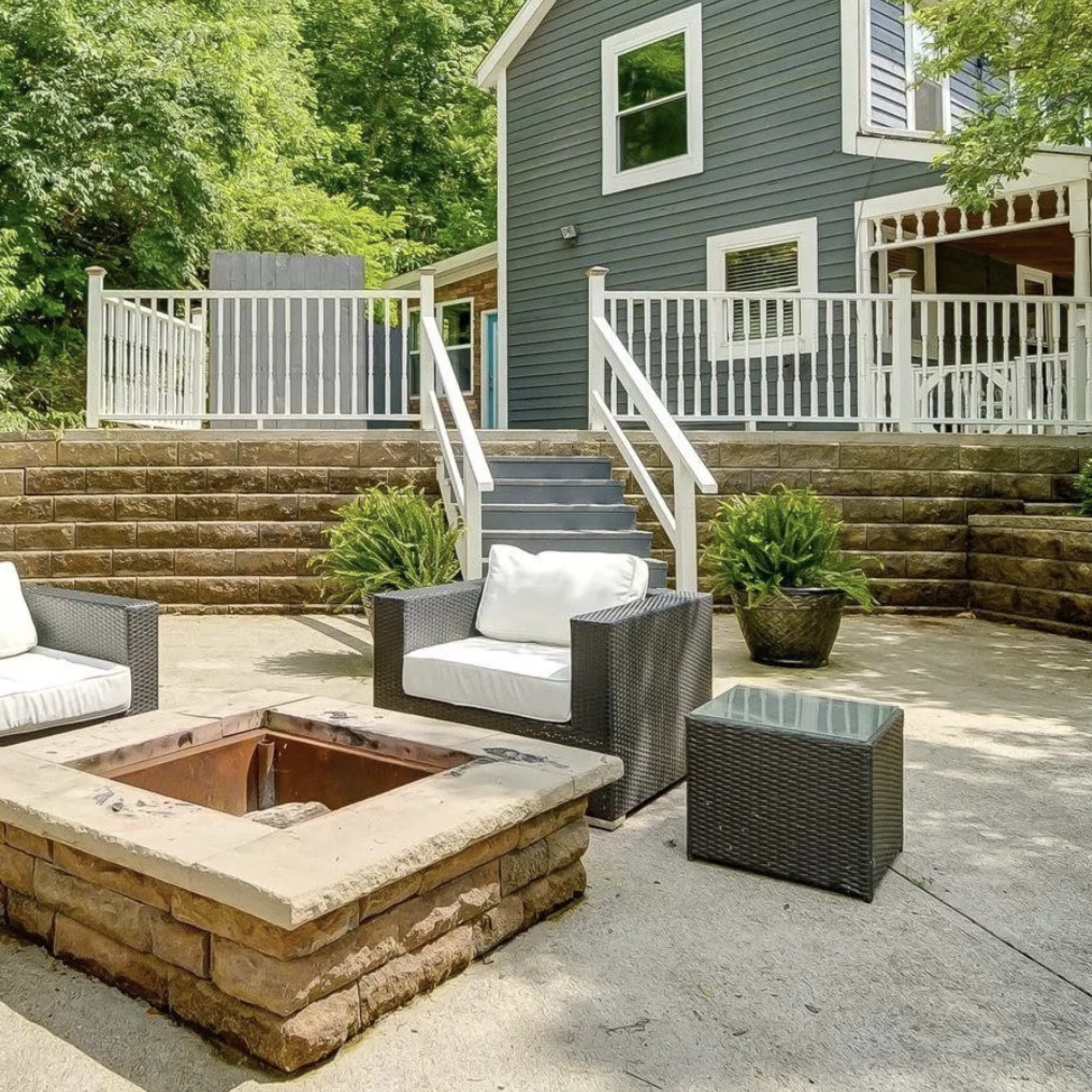 A stone fire pit is centered on a patio with two white cushioned chairs, surrounded by greenery and steps leading to a house.