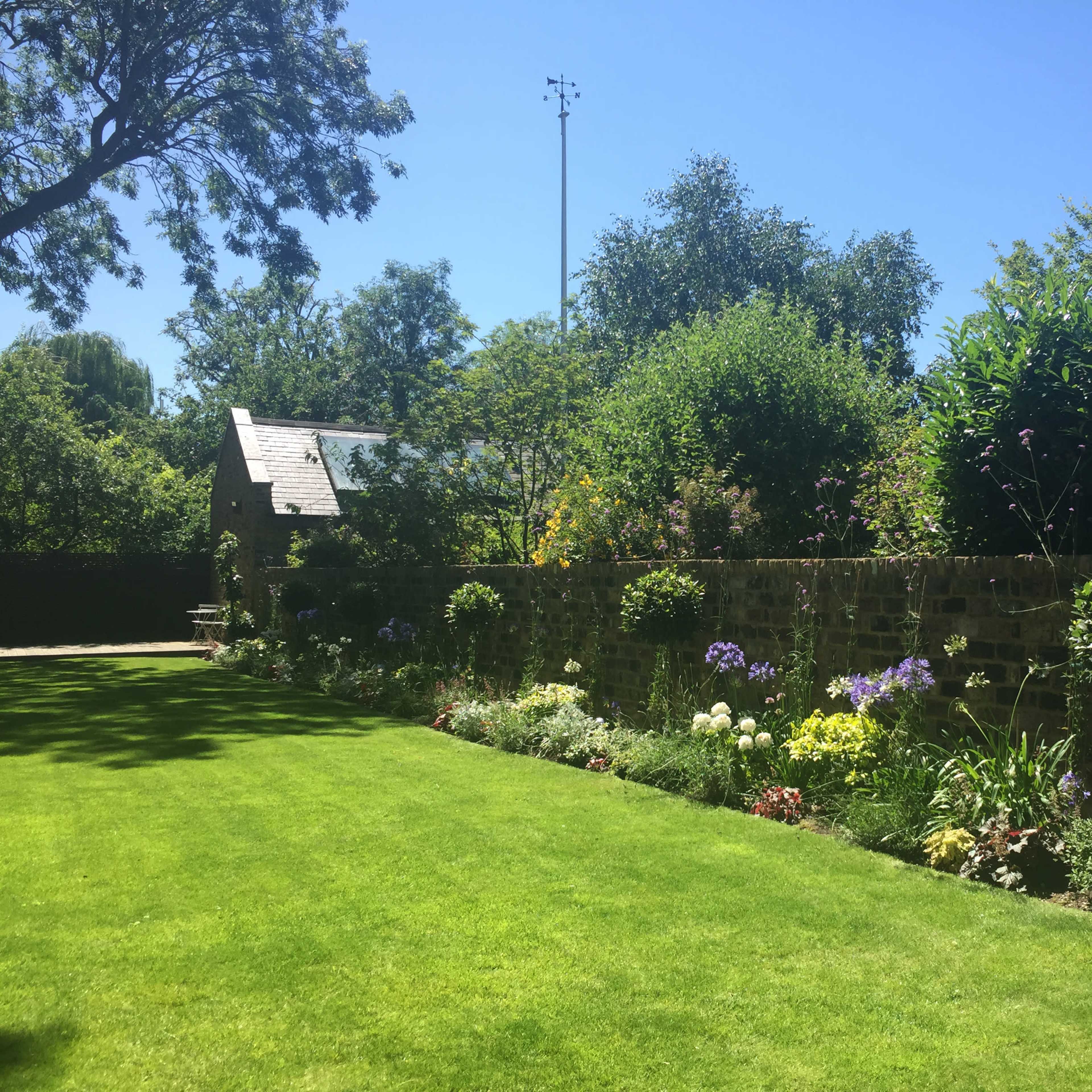 A tidy garden with a well-manicured lawn, colorful flowerbeds along a stone wall, and trees in the background under a clear blue sky.