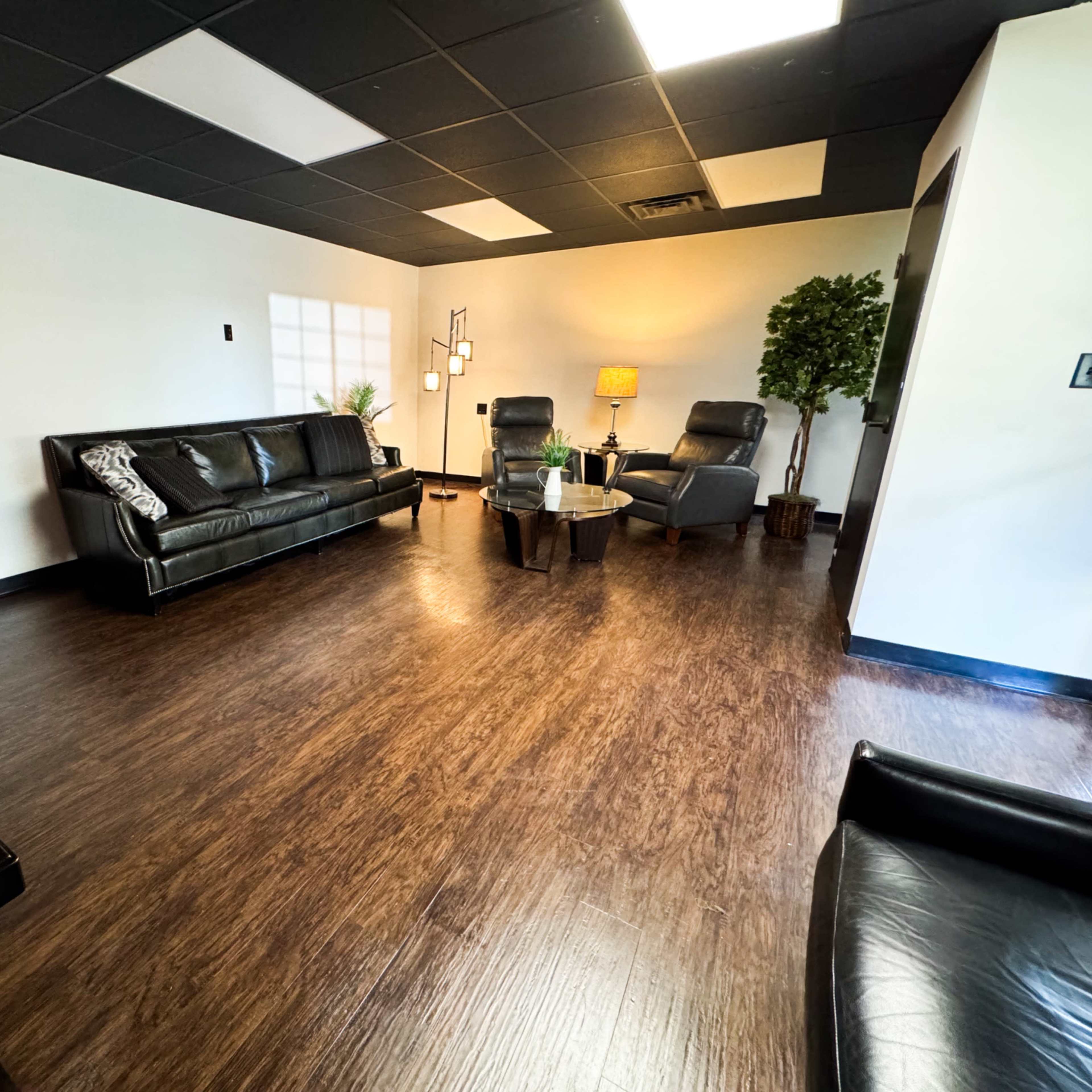 A modern, minimalist waiting room with black leather sofas, two armchairs, a coffee table, and decorative plants.