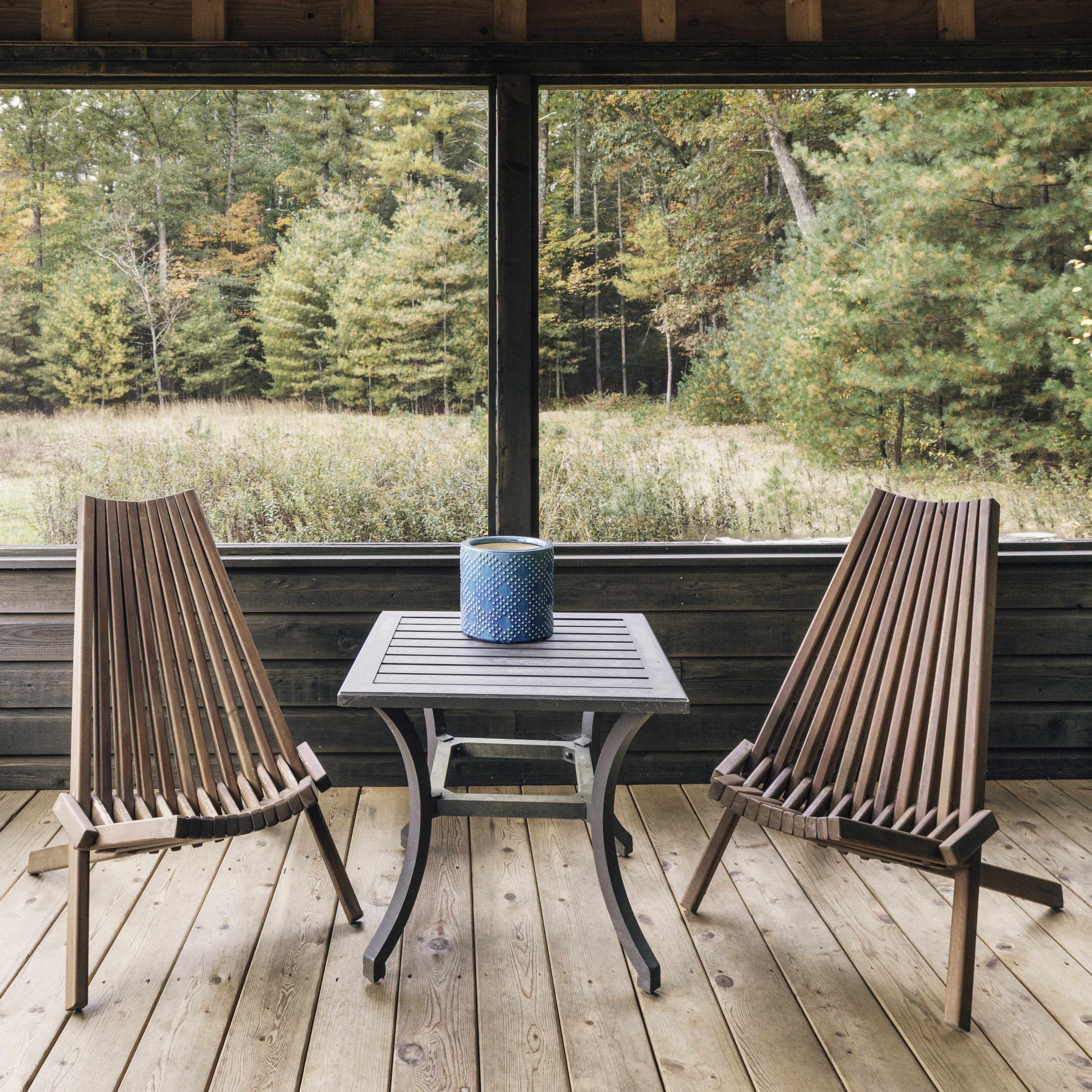 Two wooden chairs and a small table sit on a wooden porch, overlooking a forested area through a screened wall.