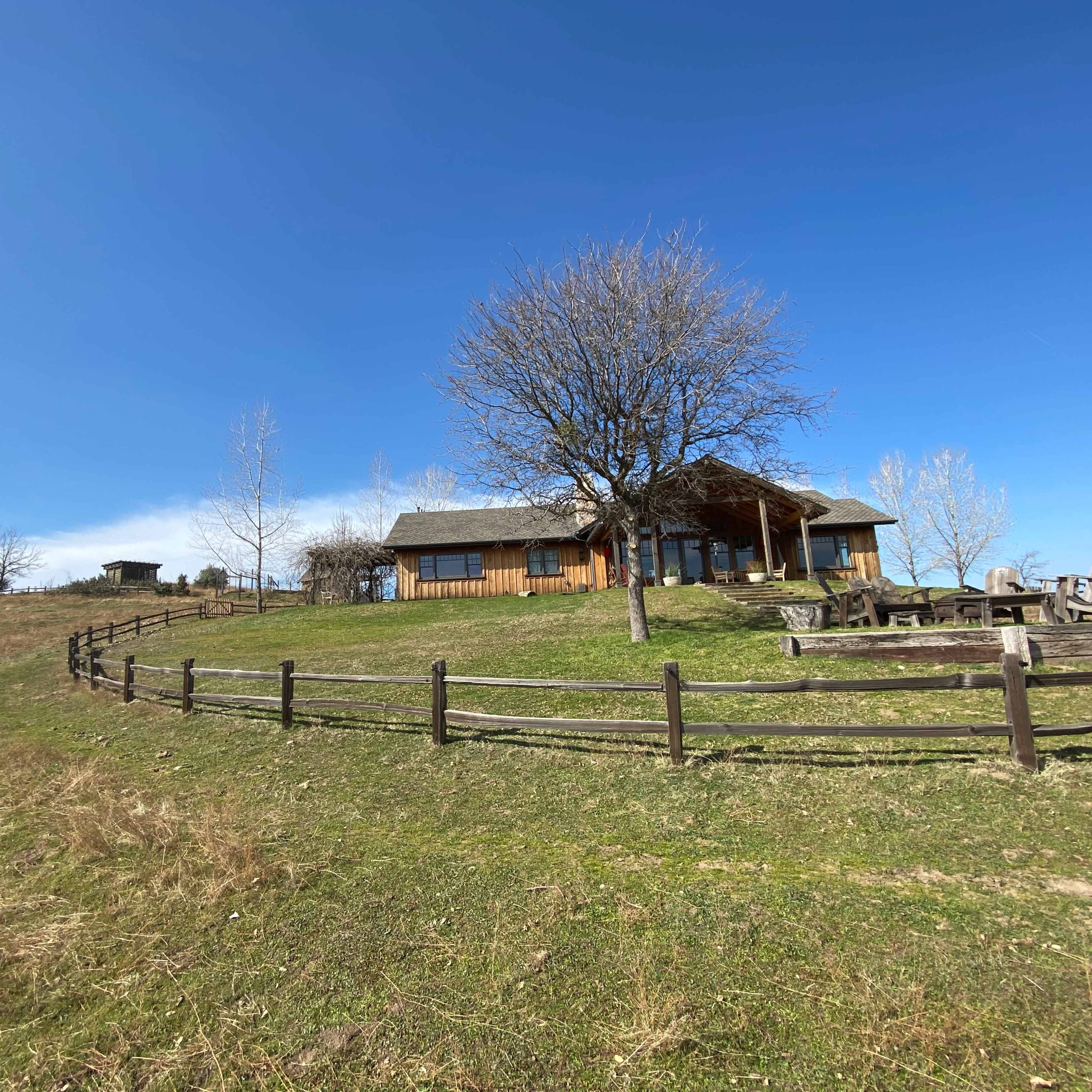 A wooden house sits on a hillside surrounded by a fence and sparse trees under a clear blue sky.