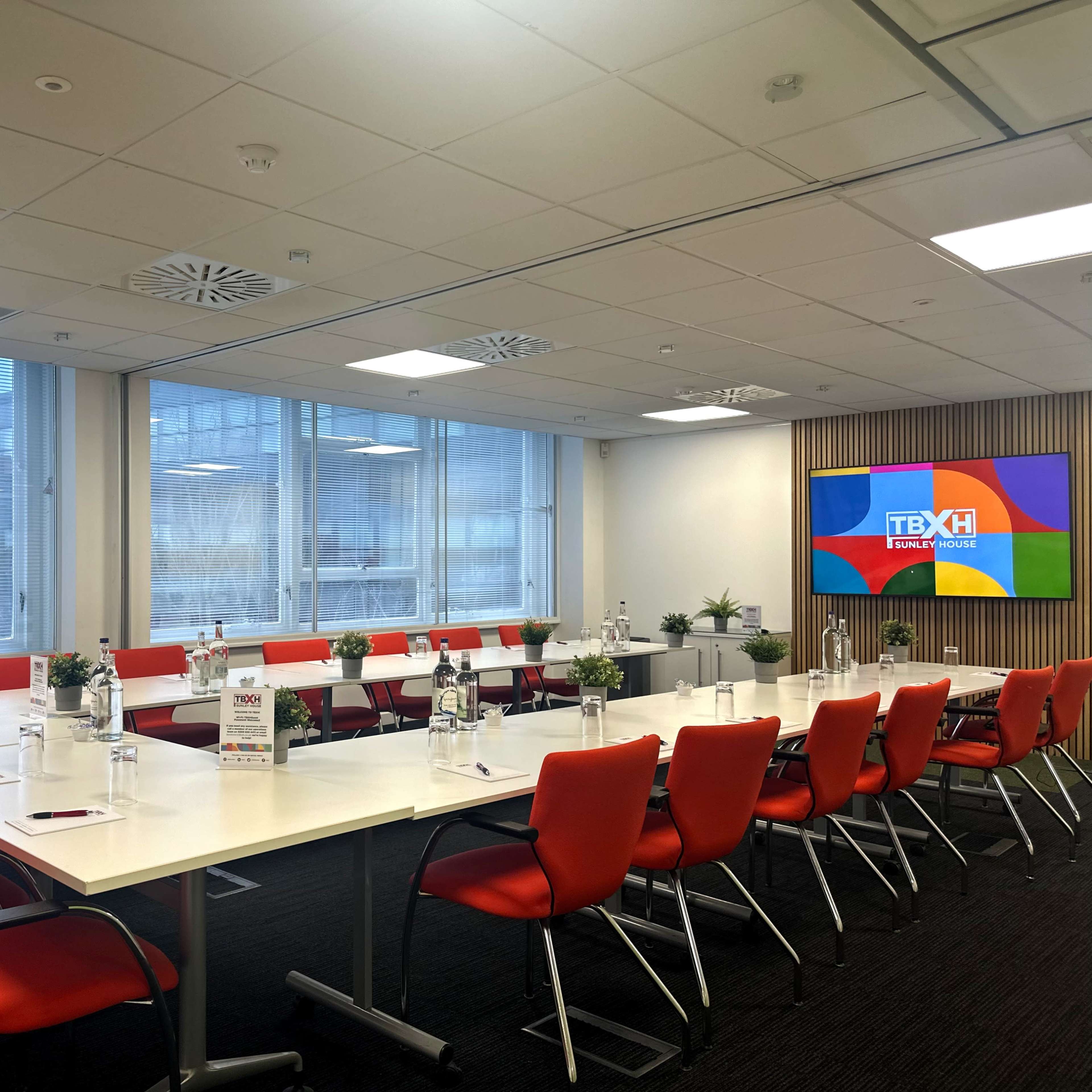 A modern conference room features a long table with red chairs, glass windows, and a large screen displaying a logo.