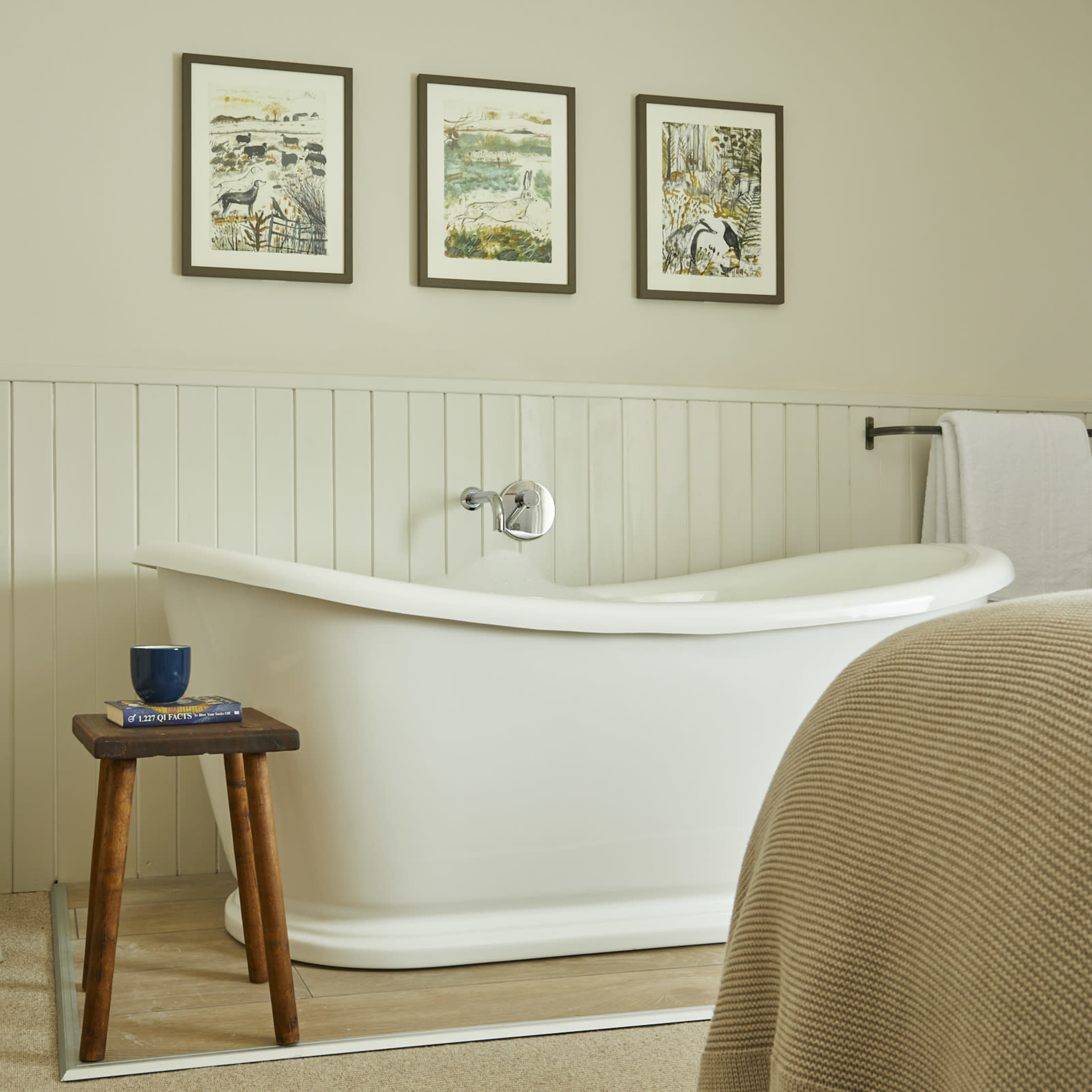 A bright bathroom featuring a white freestanding bathtub, a wicker chair, and three framed artworks on the wall.