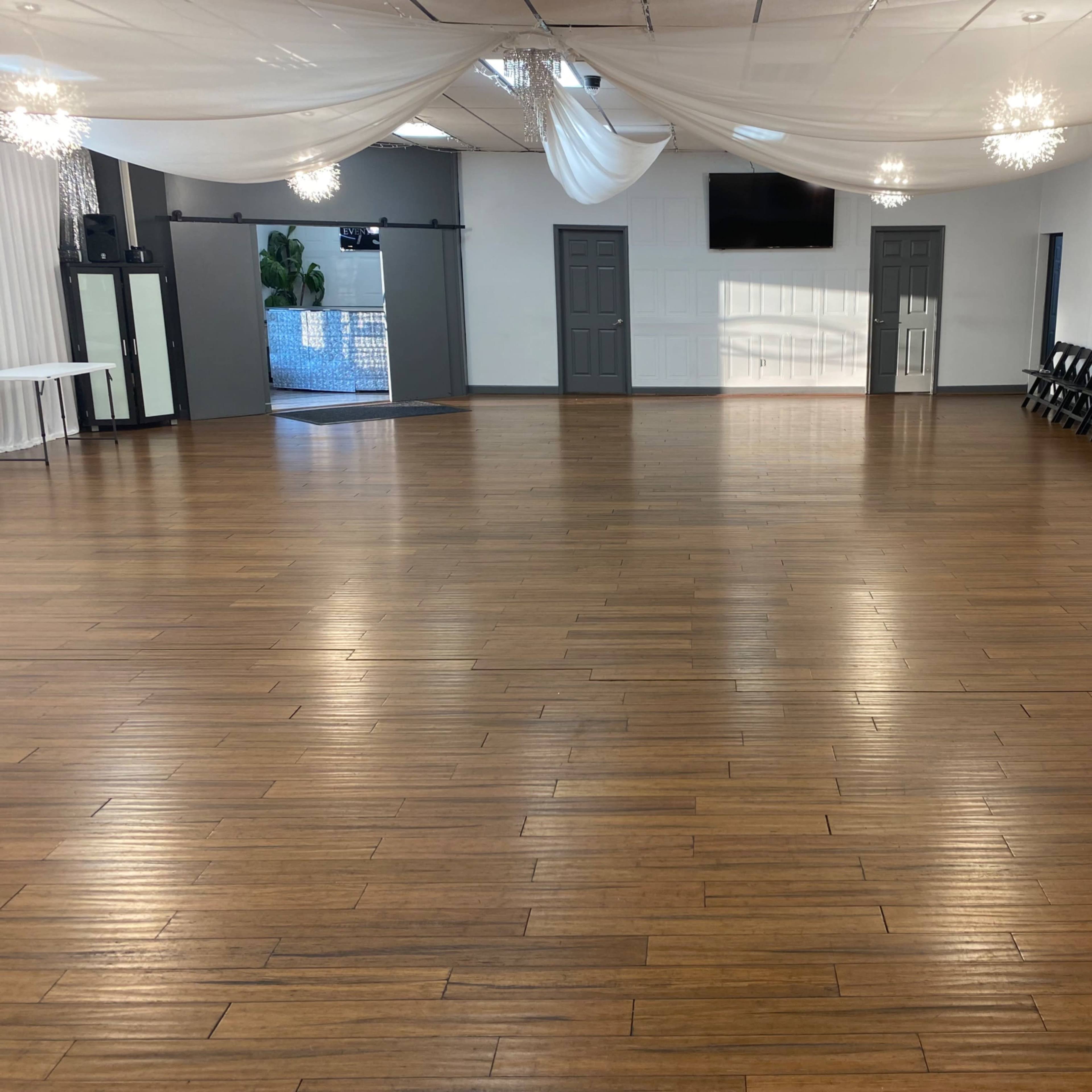 A spacious event hall with a polished wooden floor, white draped ceiling, and rows of black folding chairs against one wall.