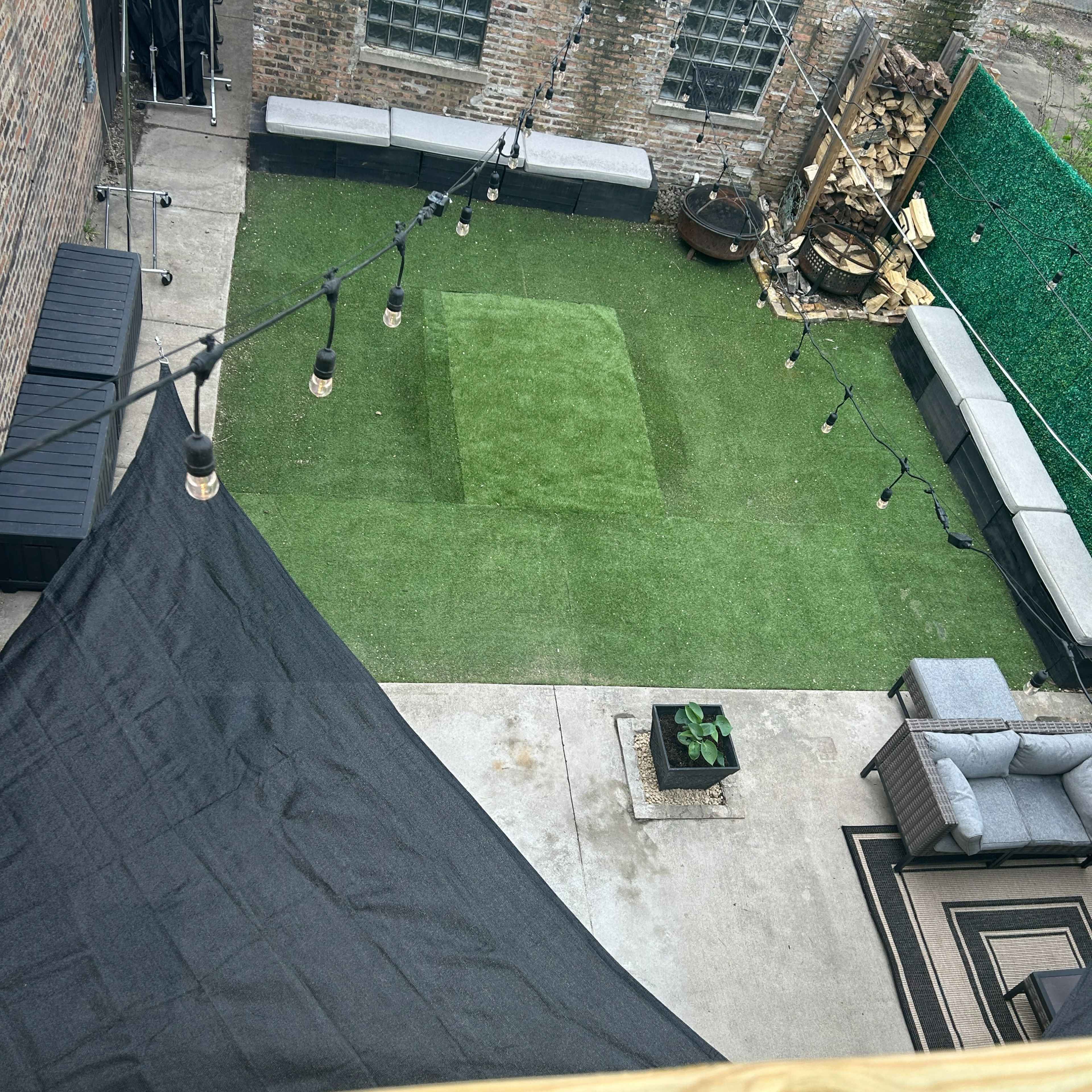 A small outdoor space featuring artificial grass, seating areas, and string lights, surrounded by brick walls and a greenery-covered backdrop.