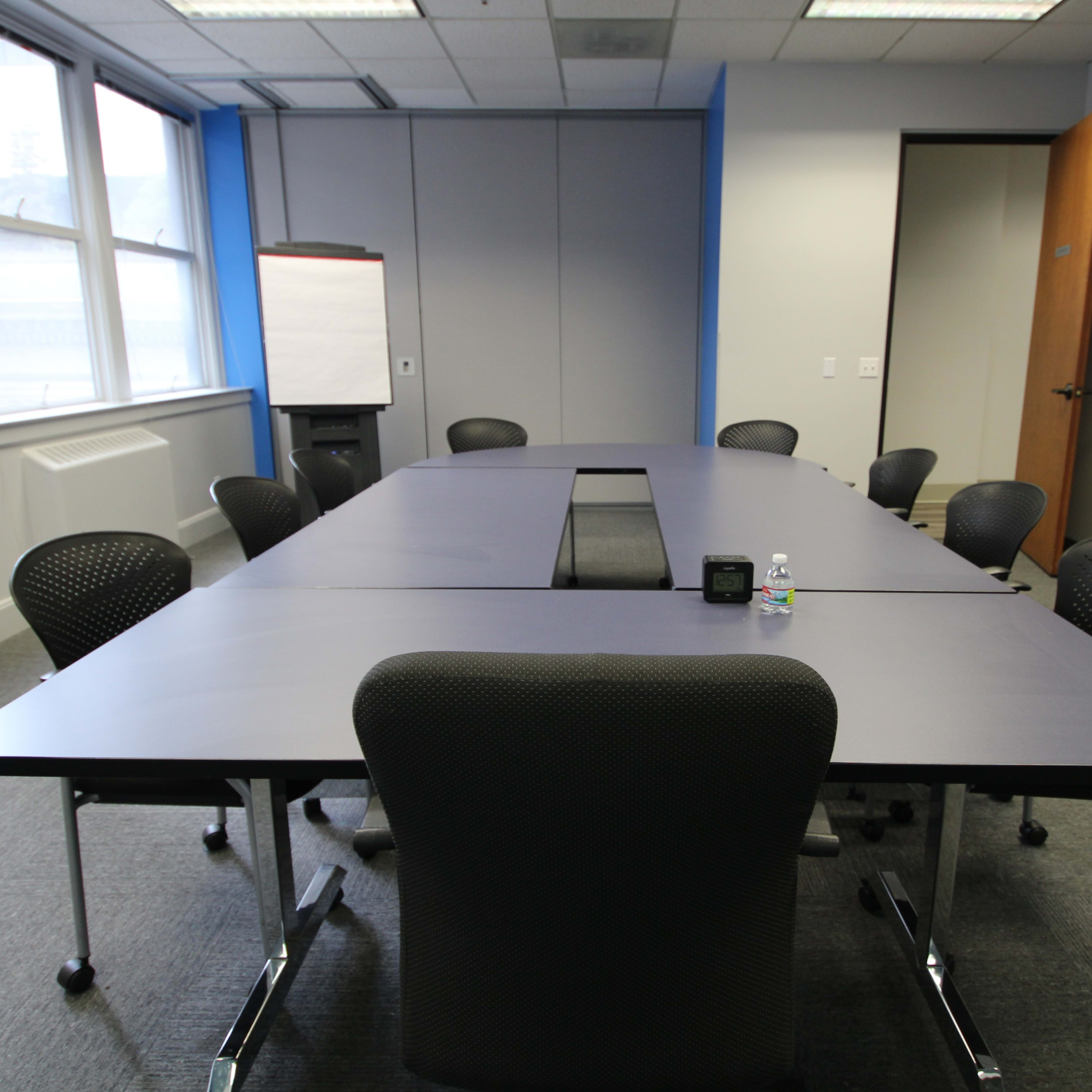 A large conference room features a long table surrounded by black chairs, with a whiteboard and a monitor on the walls.