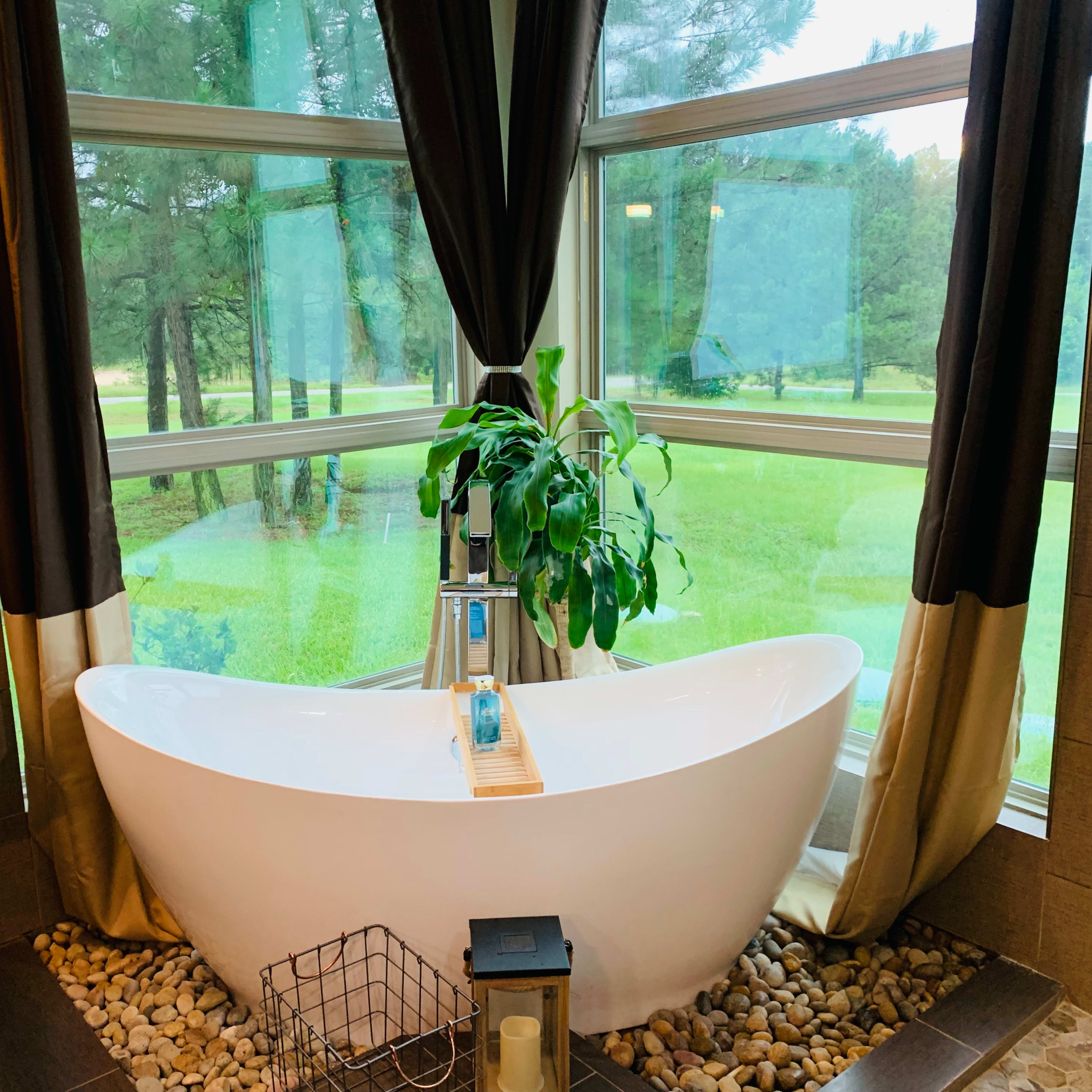 A freestanding bathtub is positioned in front of large windows that offer a view of a grassy area and trees outside.
