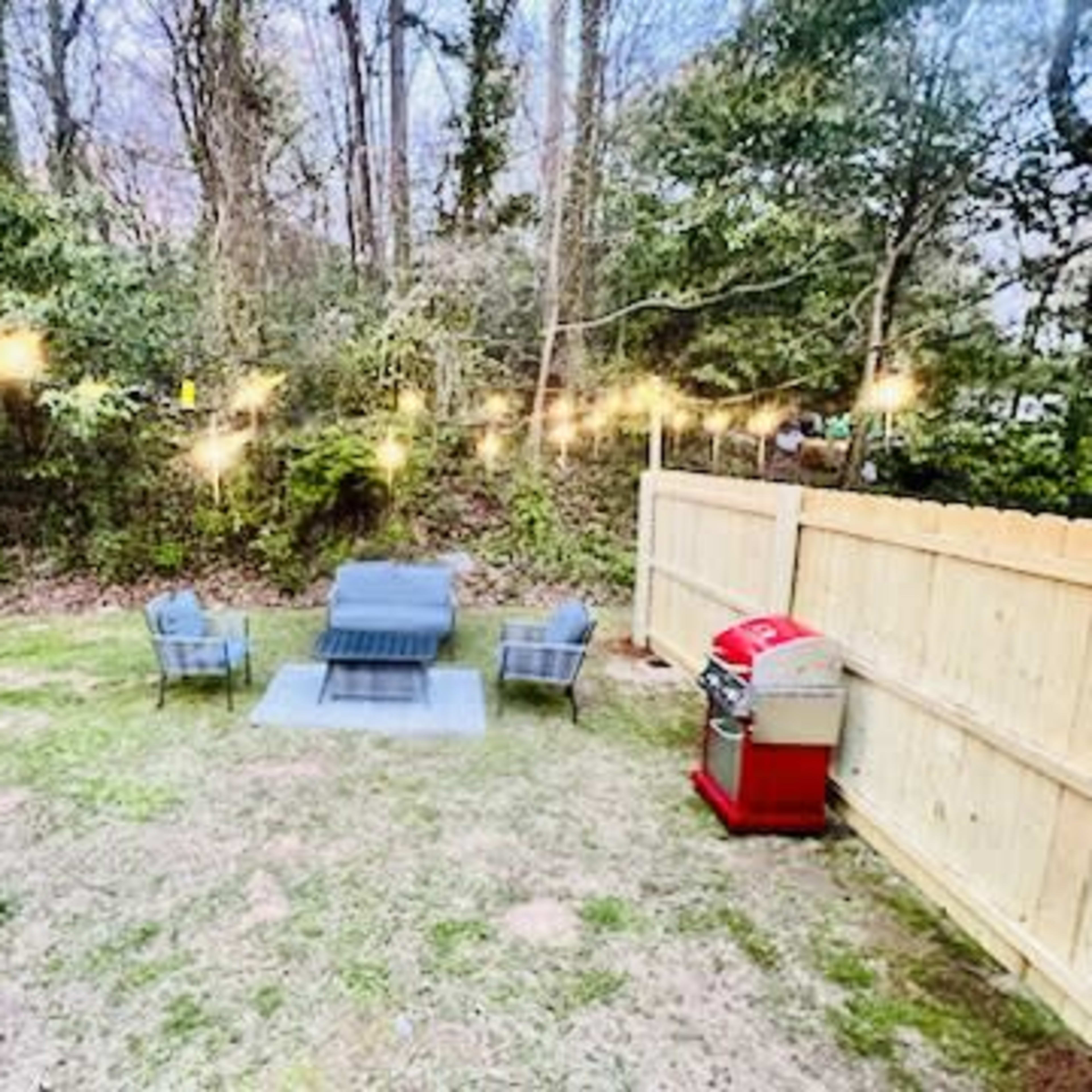 A backyard setup with four chairs arranged around a fire pit, string lights overhead, and a red grill placed against a wooden fence.