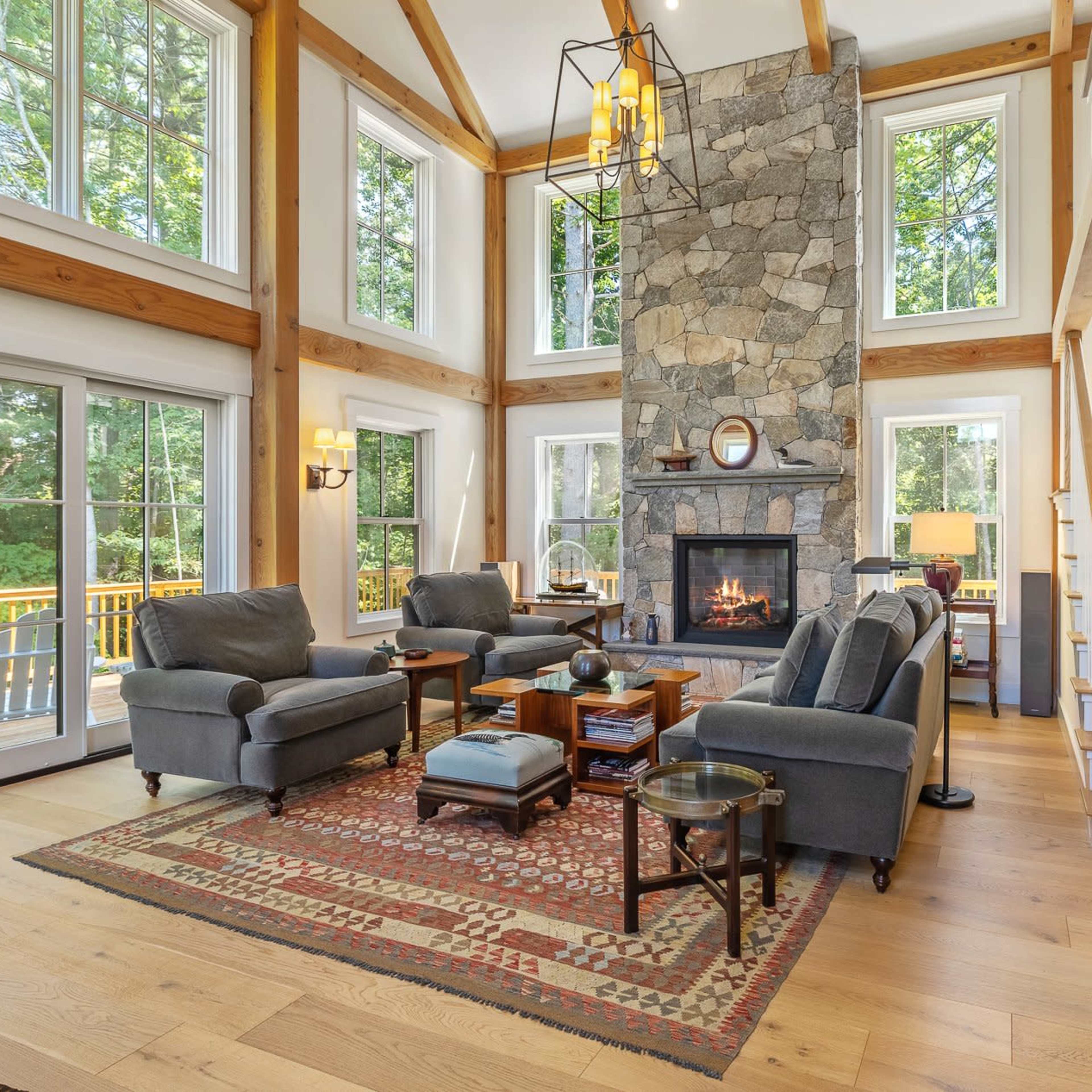 A spacious living room features a stone fireplace, wooden beams, large windows with views of the outdoors, and comfortable seating arrangements.