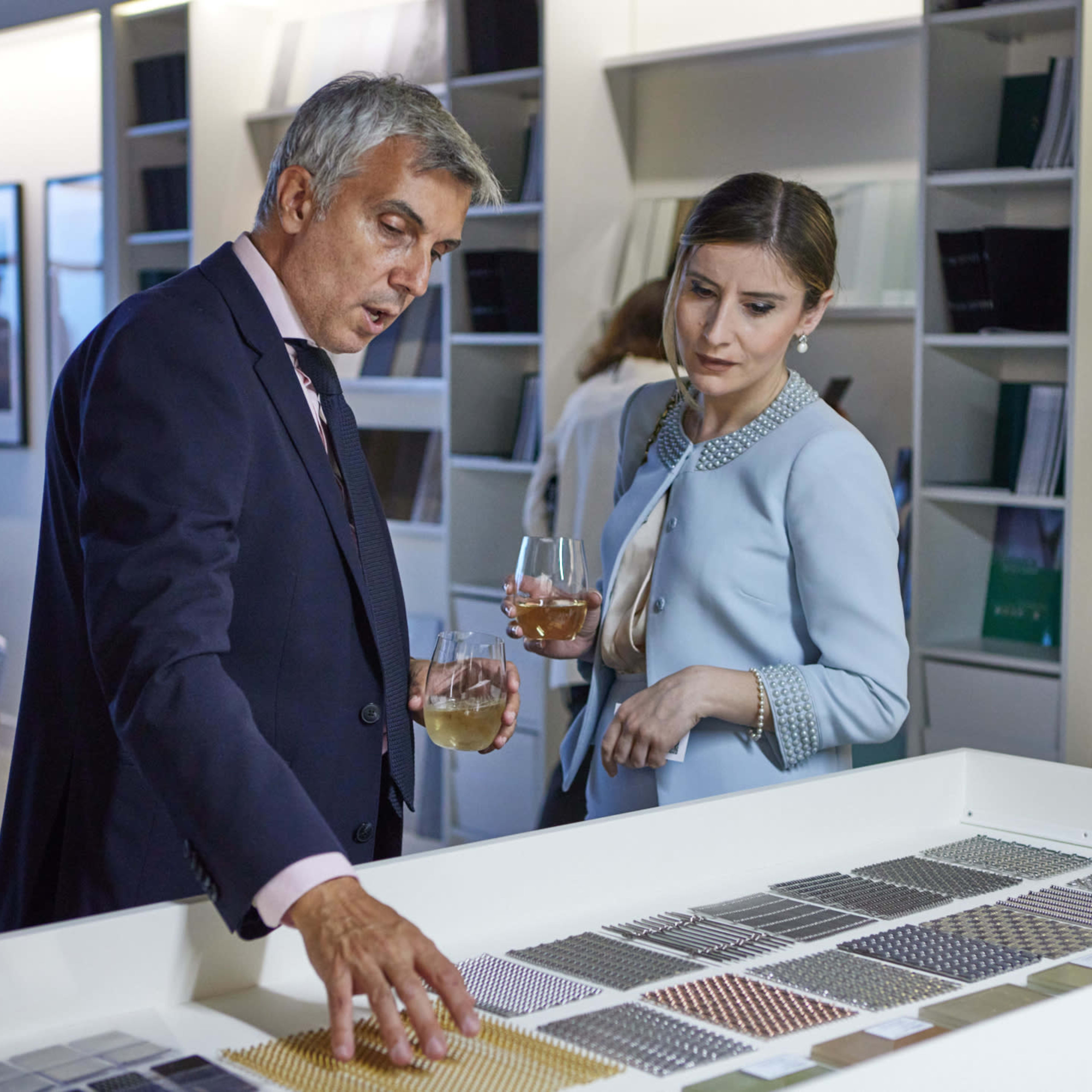 A man and a woman stand by a display of material samples, discussing options while holding glasses of drinks.