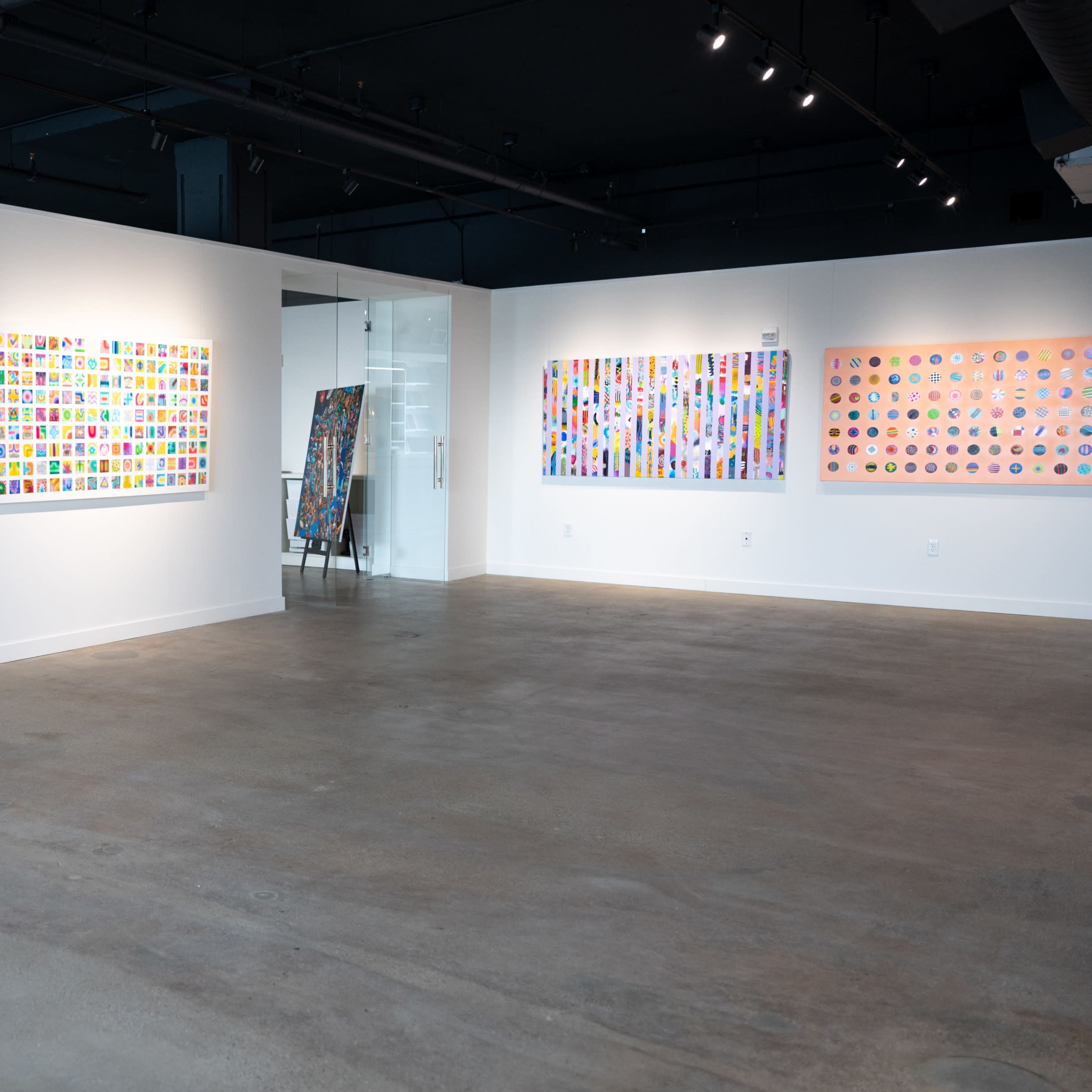 An art gallery features three vibrant abstract paintings displayed on white walls, with a spacious concrete floor and modern lighting.