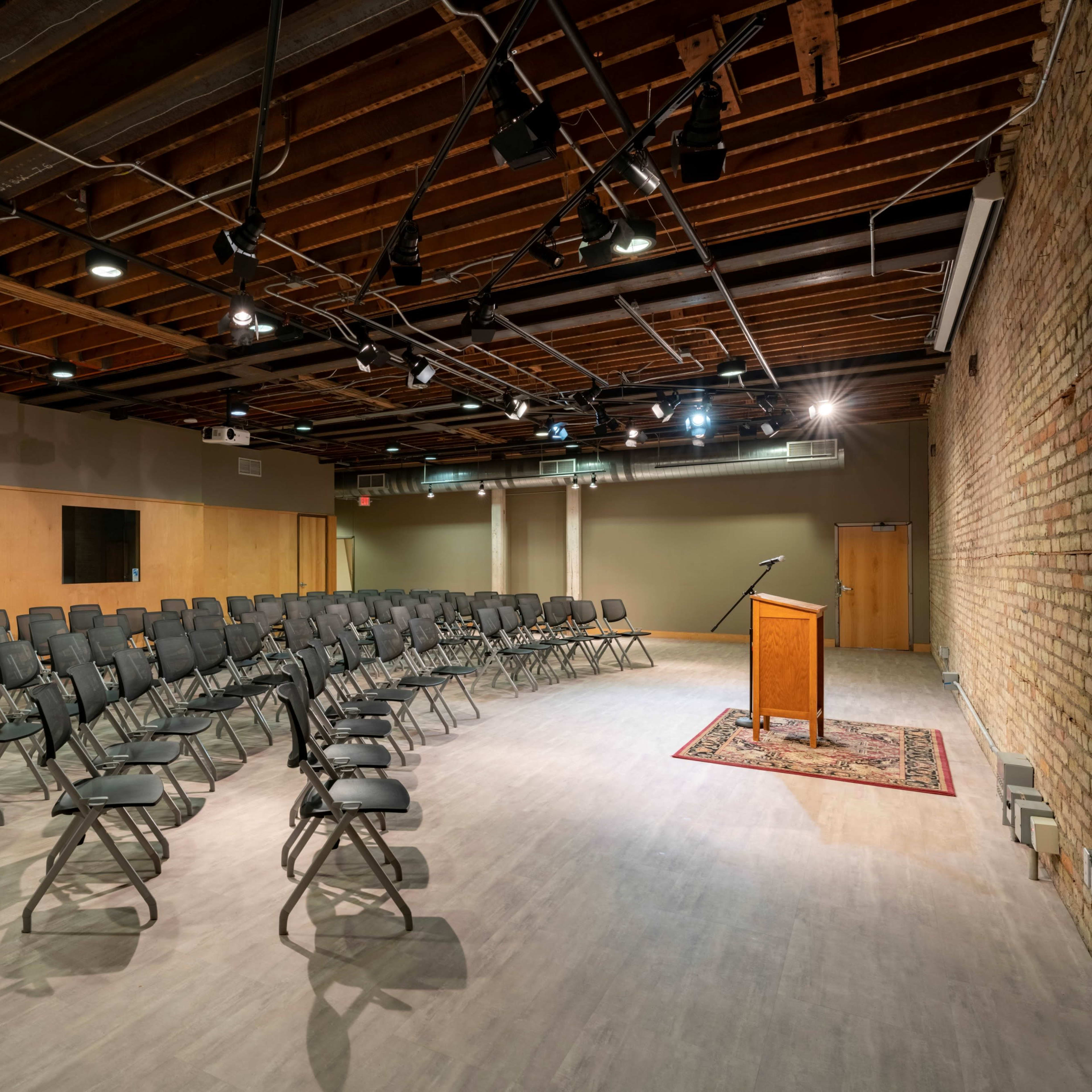 A spacious room with rows of black folding chairs arranged facing a wooden podium on a rug, with exposed brick walls and overhead lighting.