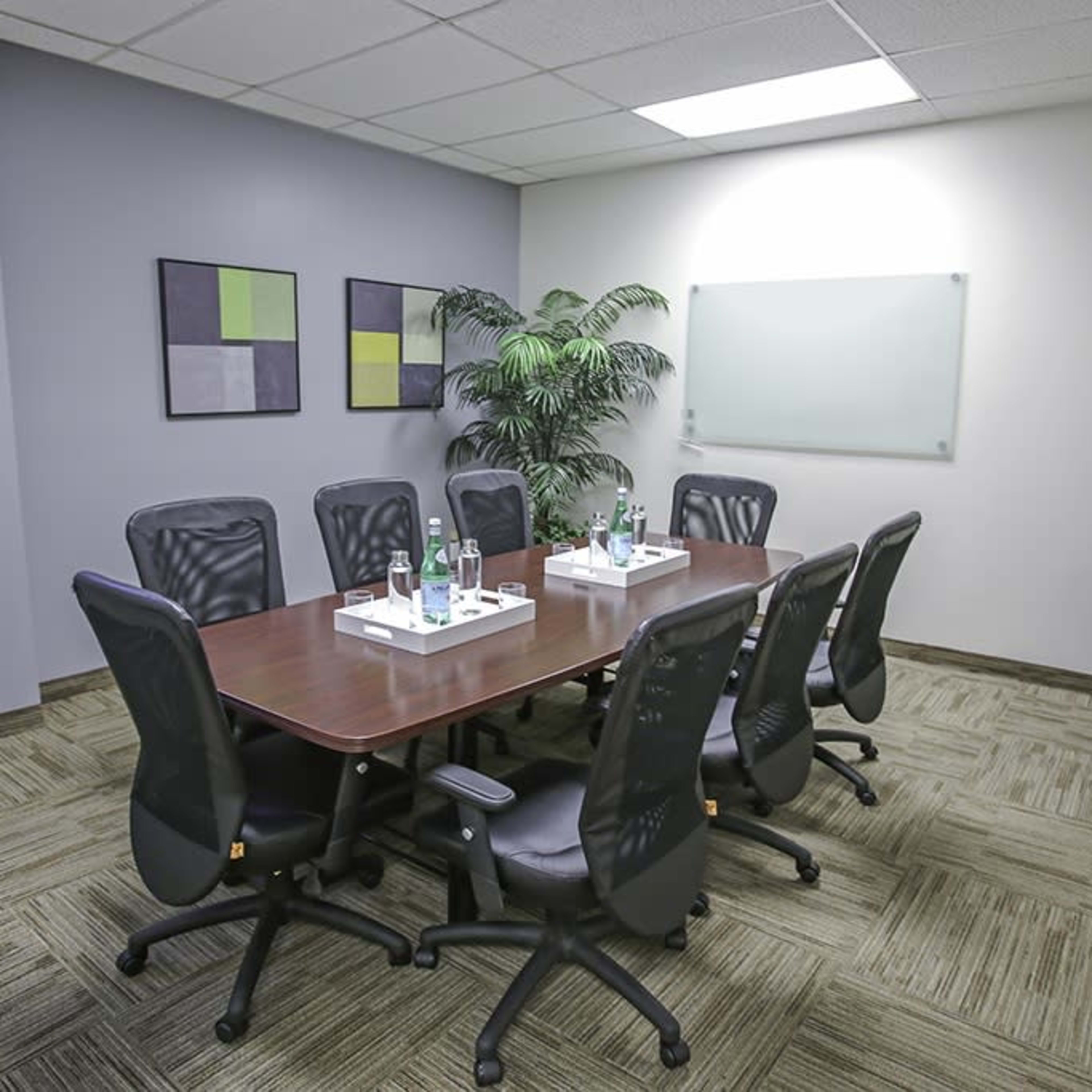 A conference room features a large wooden table surrounded by eight black chairs, with a plant in the corner and artwork on the wall.