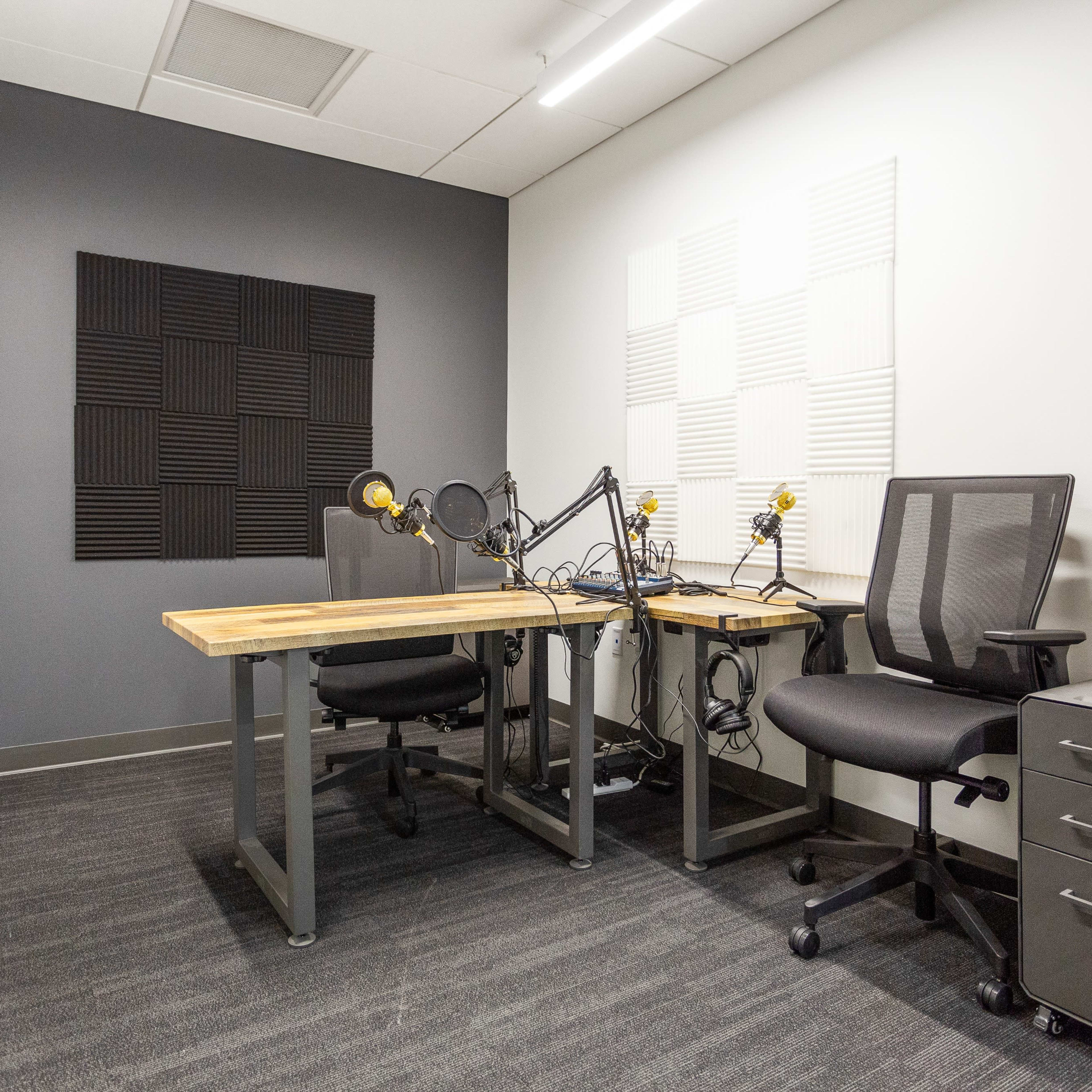 A small recording studio with two microphone setups on wooden tables, black chairs, and soundproofing panels on the walls.