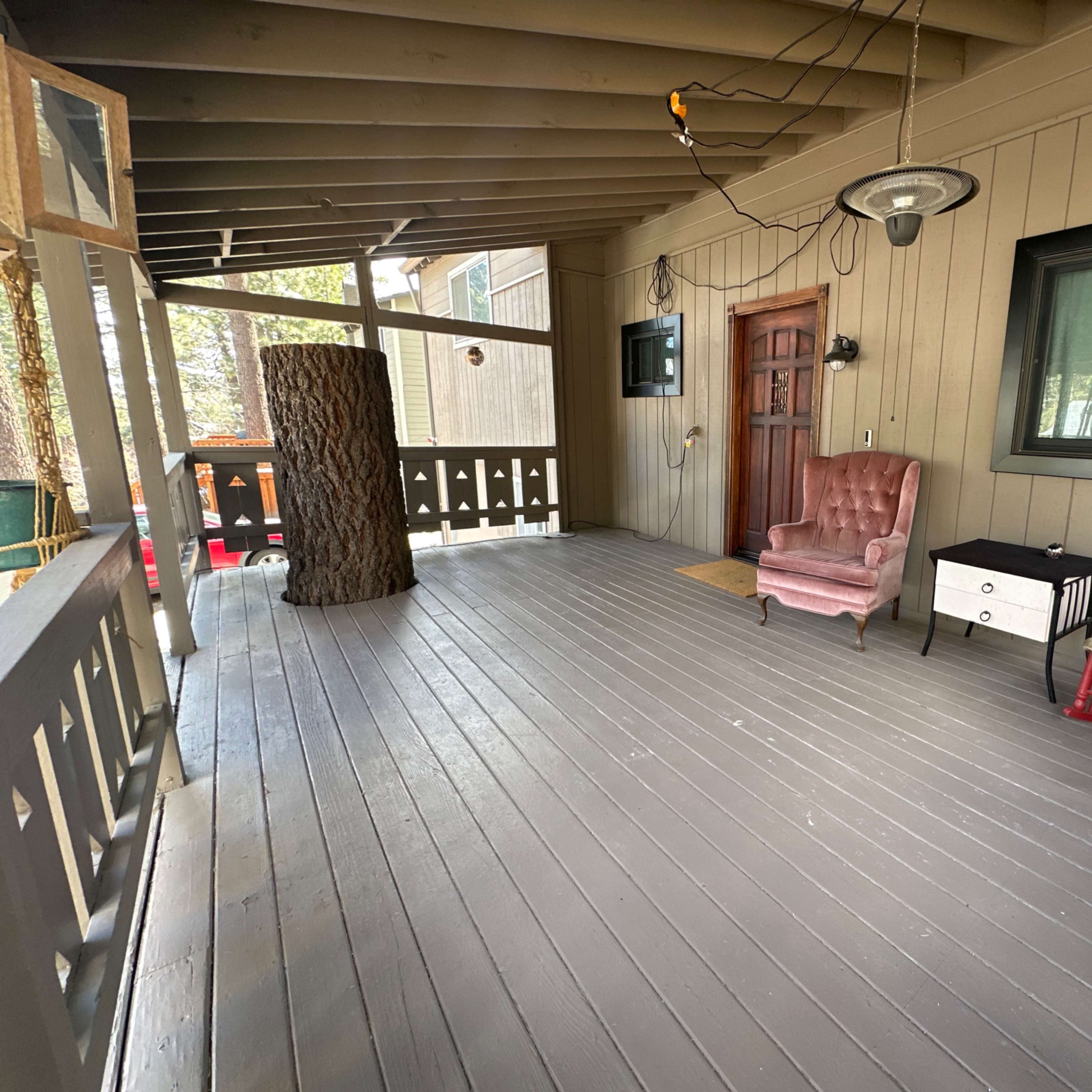 A covered porch with wooden flooring, featuring a rustic tree trunk, a pink armchair, a small black and white table, and a red rocking chair against a backdrop of wooden walls and a window.
