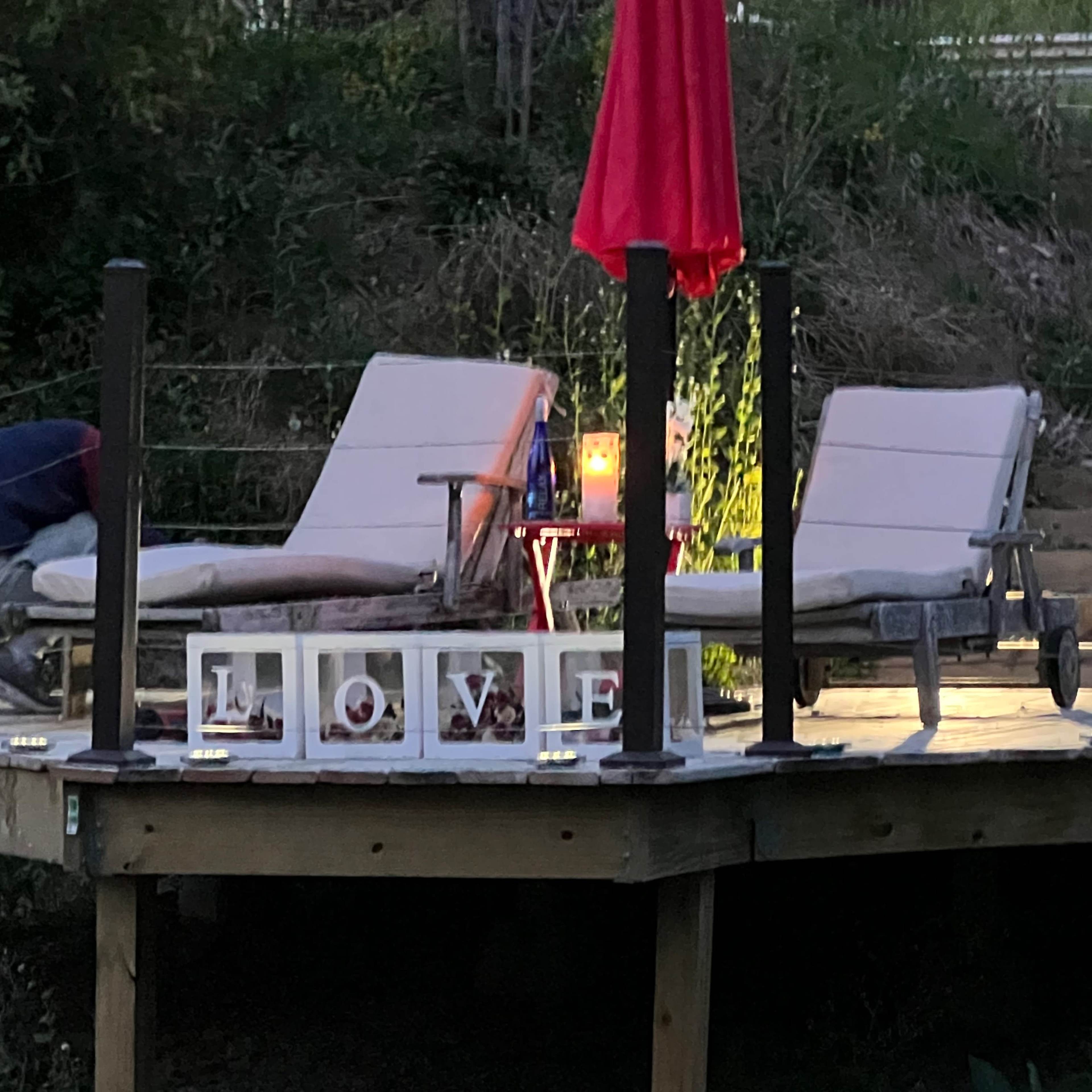 A person is adjusting a lounge chair on a wooden deck illuminated by a lamp and surrounded by decorative letters spelling "LOVE."
