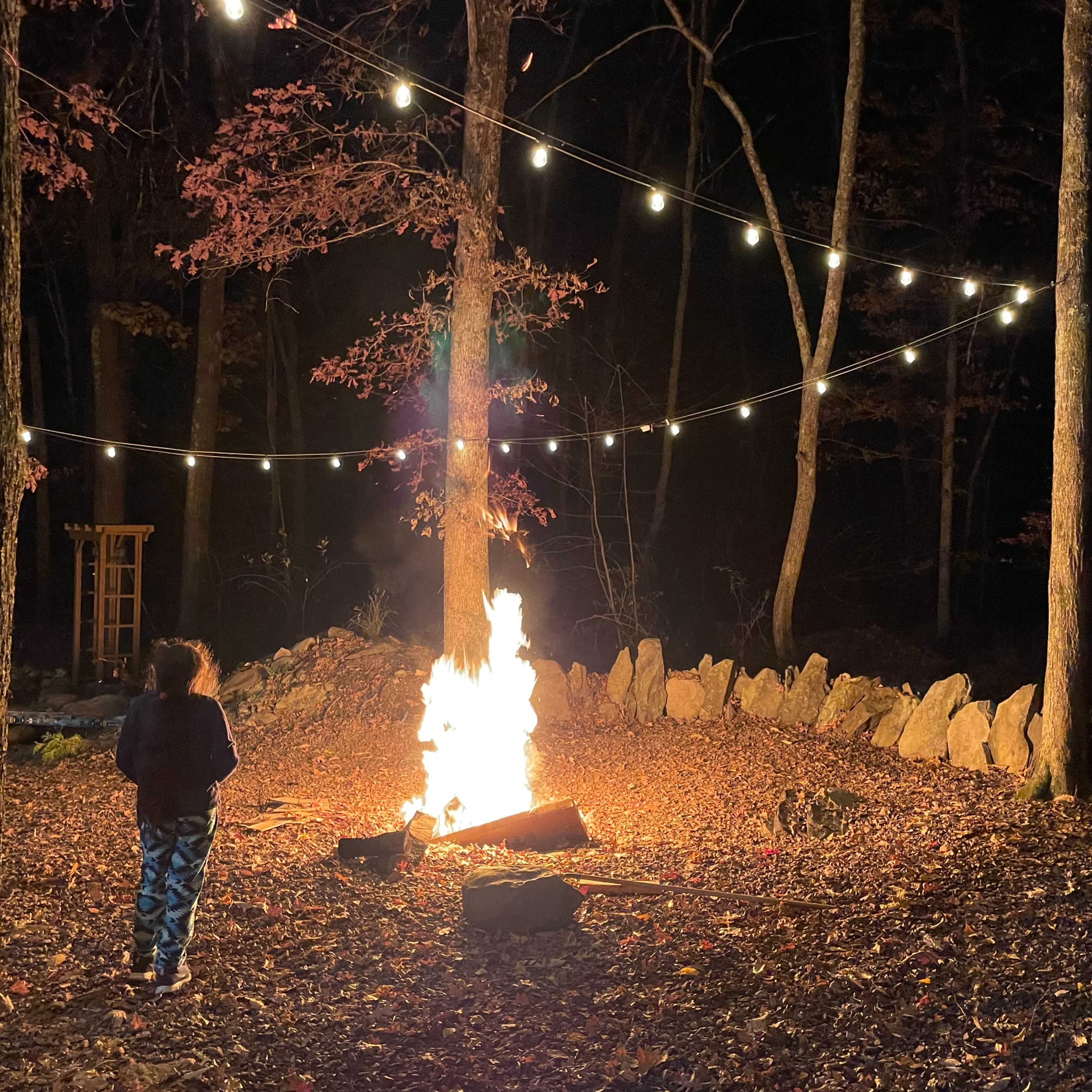 A person stands near a fire surrounded by trees, with string lights hanging overhead in a wooded area at night.