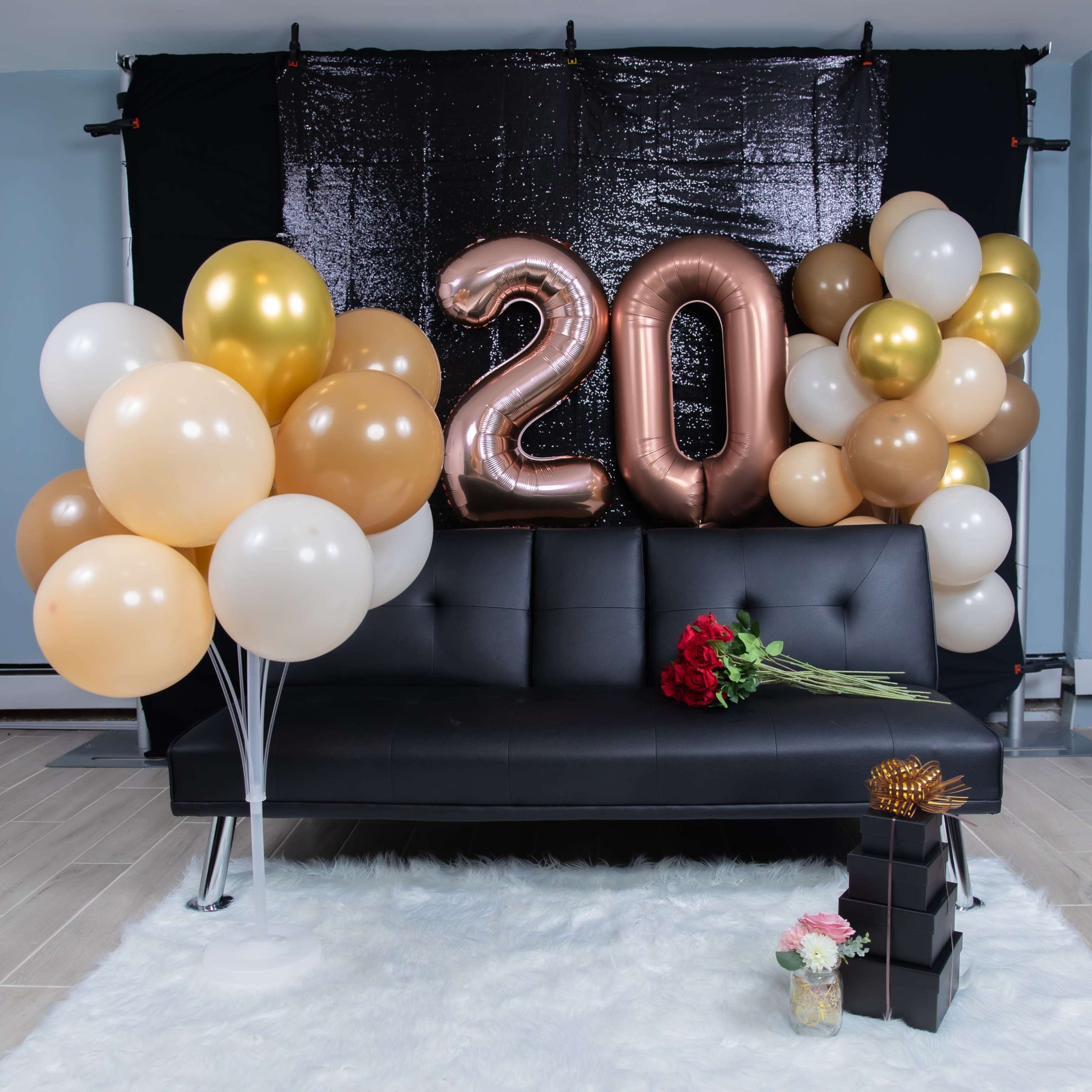 A black sofa is decorated with large metallic "20" balloons and an assortment of pastel balloons, alongside a bouquet of red roses and gift boxes.