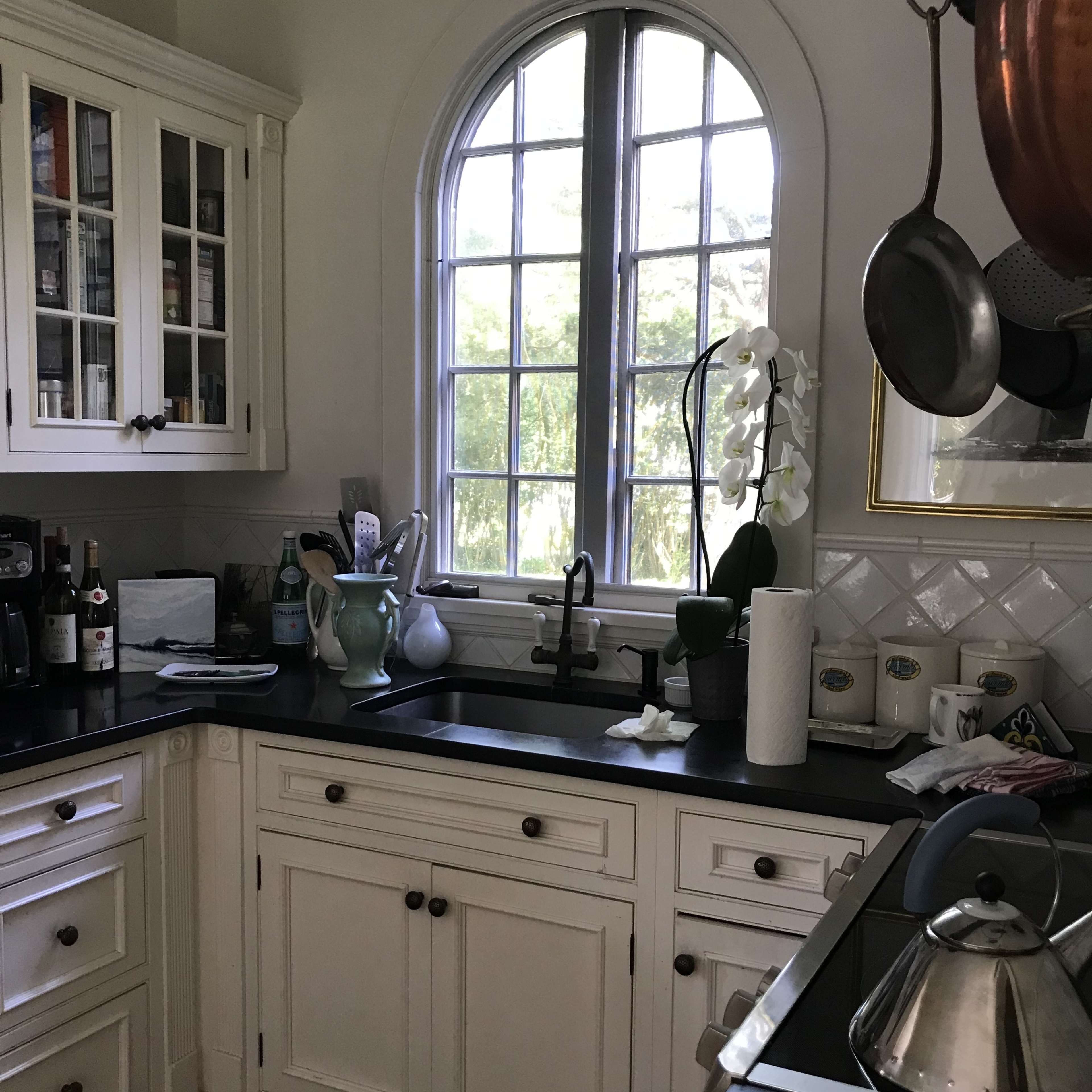 A bright kitchen with white cabinets, a black countertop, and a window featuring an arched design, overlooking greenery.